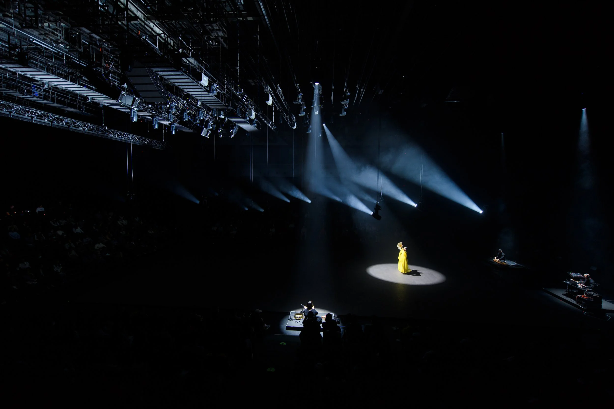A person in a bright yellow outfit stands on an empty stage under a spotlight, surrounded by darkness. Stage lighting equipment is visible overhead.