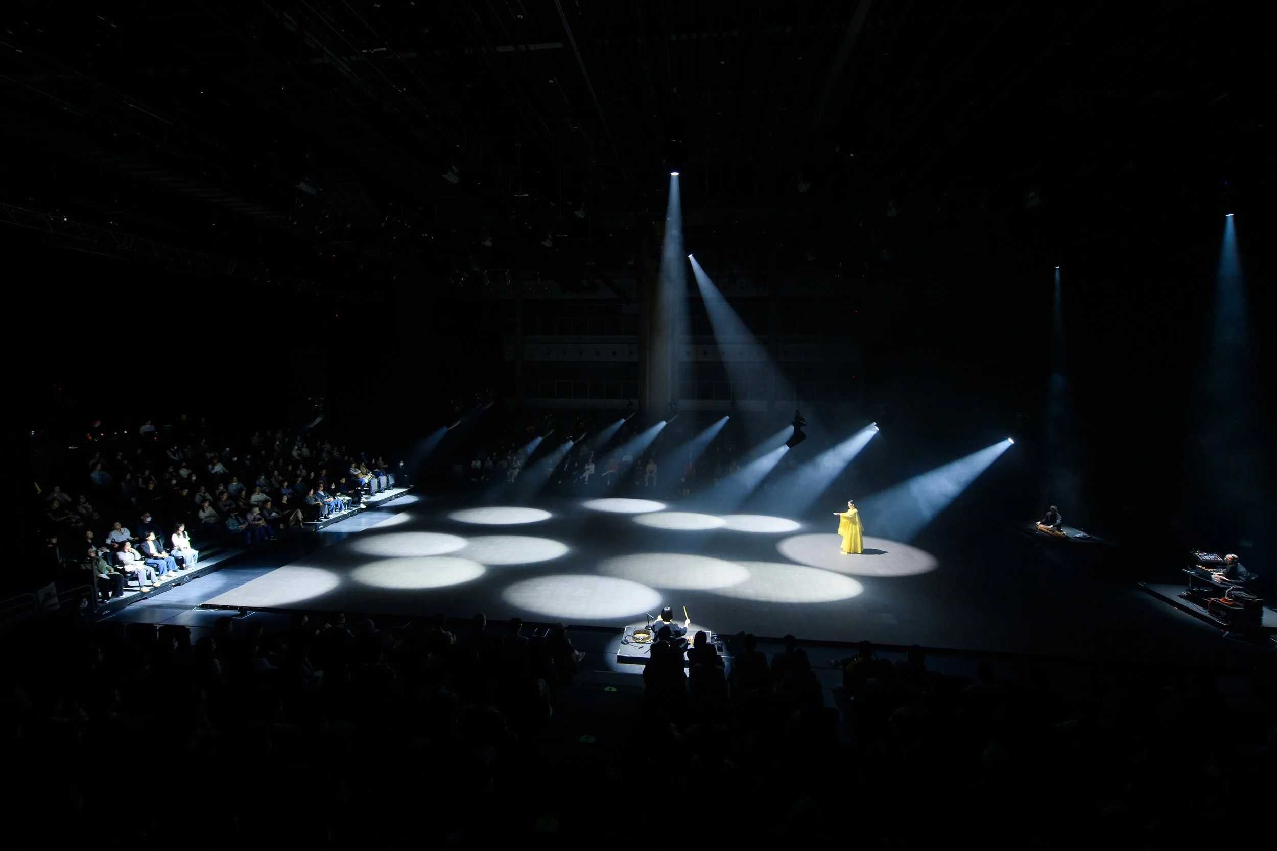 Person in yellow costume standing under spotlight on stage, surrounded by audience in dimly lit theater with multiple spotlights.