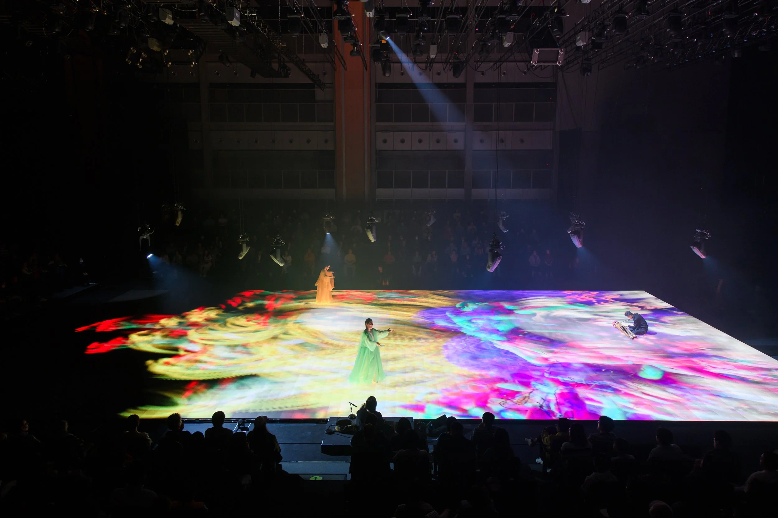 Dancers performing on a stage with colorful, digital projections and an audience watching.