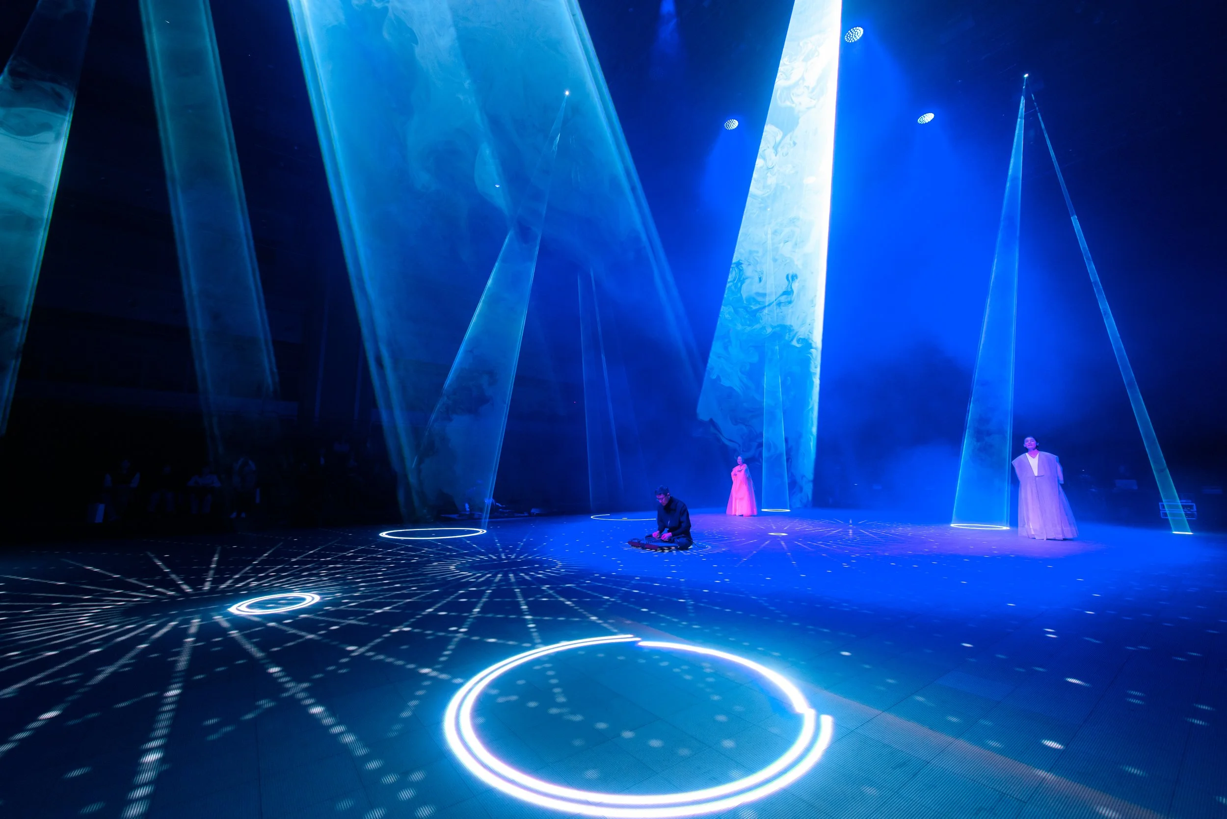 Stage performance with blue lighting and laser effects, featuring three people in robes, creating a futuristic atmosphere.
