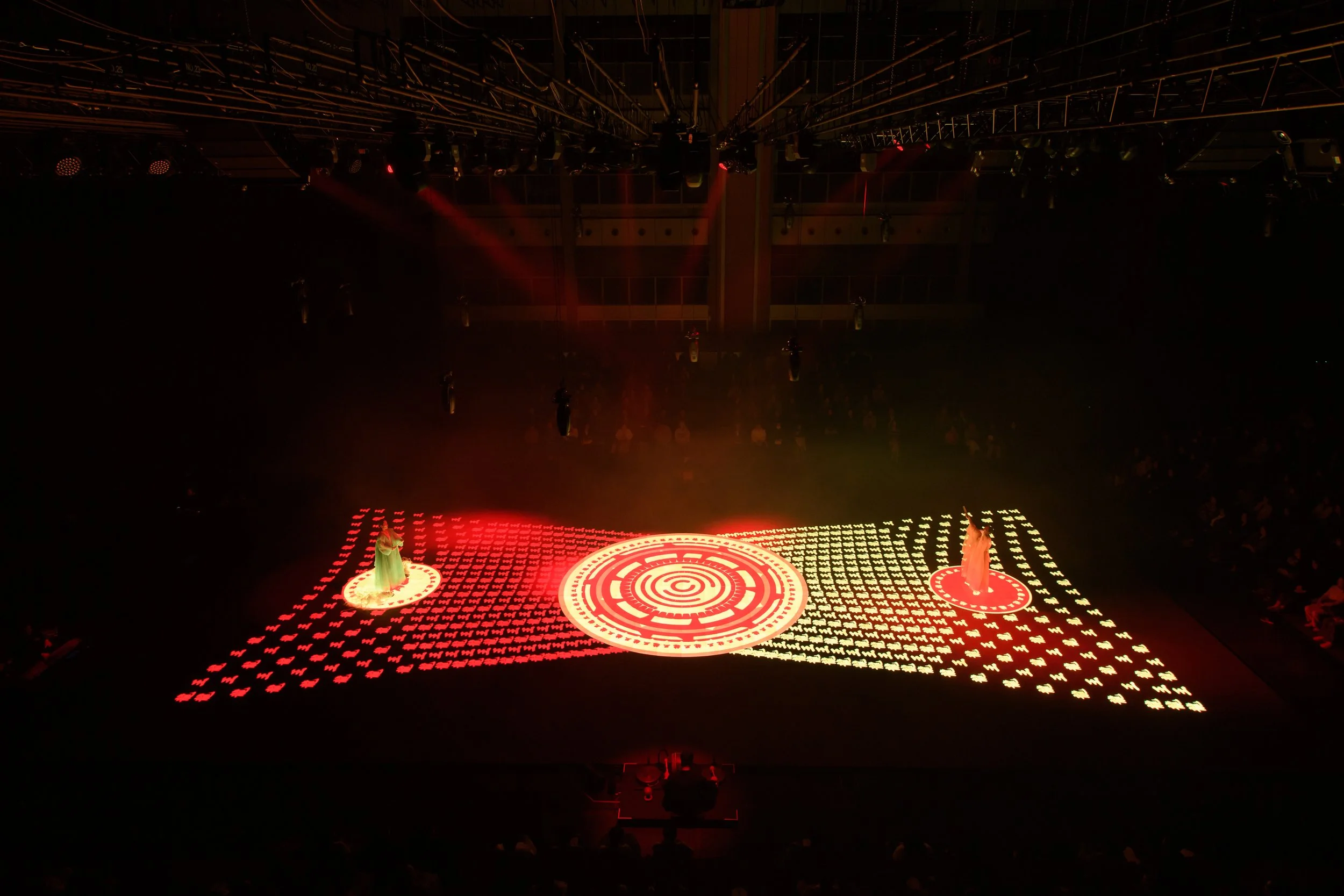 Dark stage with illuminated red and orange circle patterns, two performers in robes standing on individual spots, surrounded by a grid of light patterns.