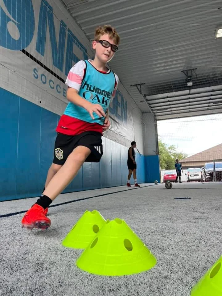 A young boy wearing glasses, a blue and white sports shirt, red shorts, and red sports shoes is practicing soccer drills with yellow cone markers inside a covered sports facility.