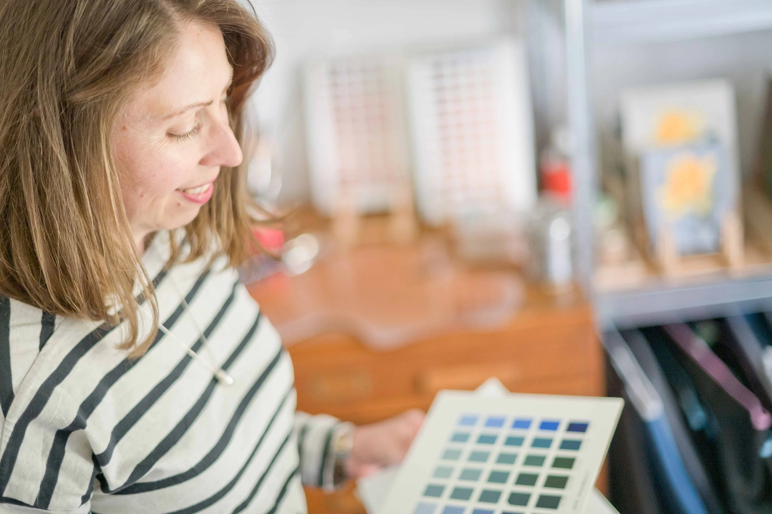 Woman with shoulder-length red hair wearing a striped shirt, smiling and holding a color palette in a workspace with blurred shelves and framed pictures in the background.