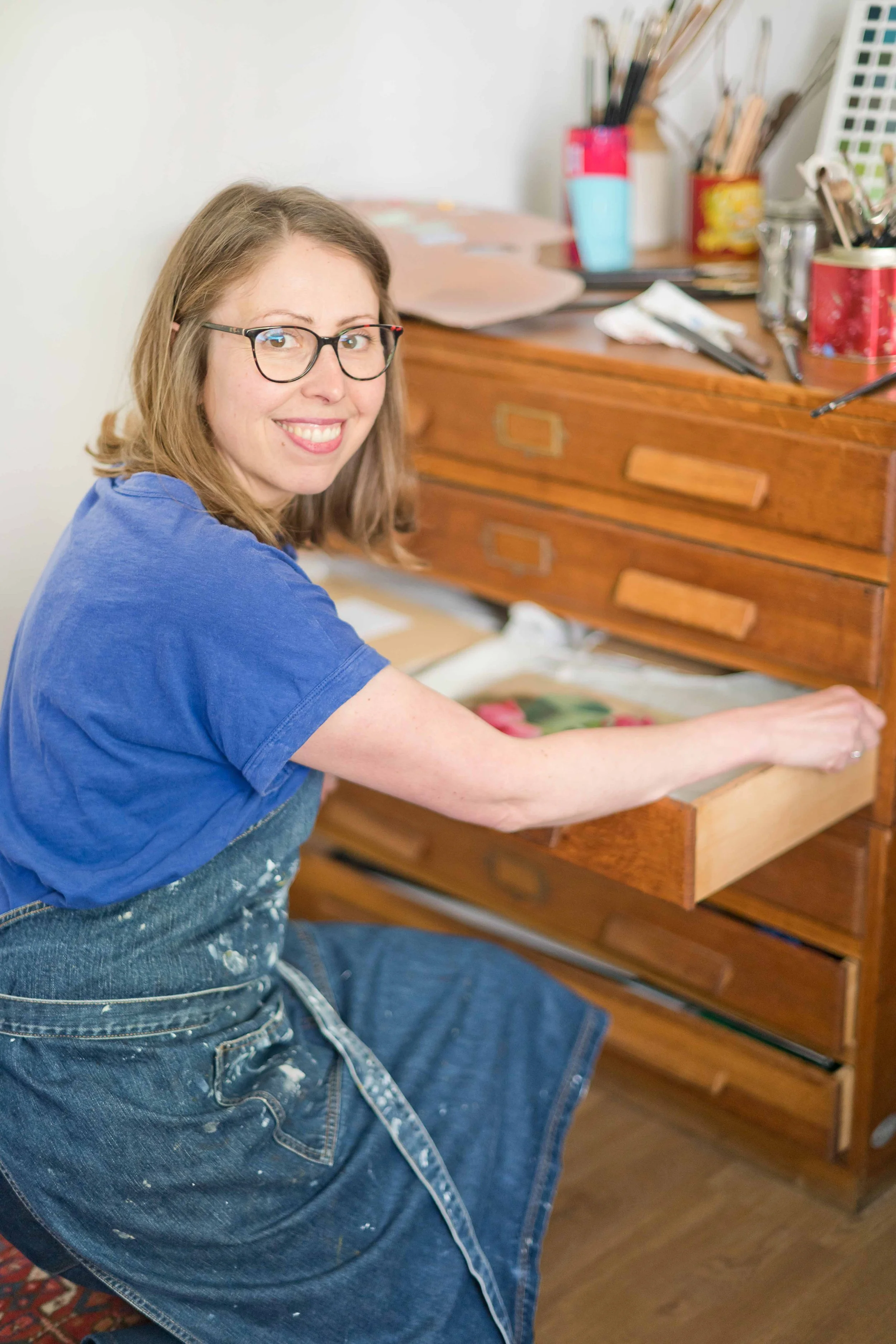 Woman wearing glasses and a blue shirt opening a wooden drawer in an art studio, with art supplies on a desk behind her.