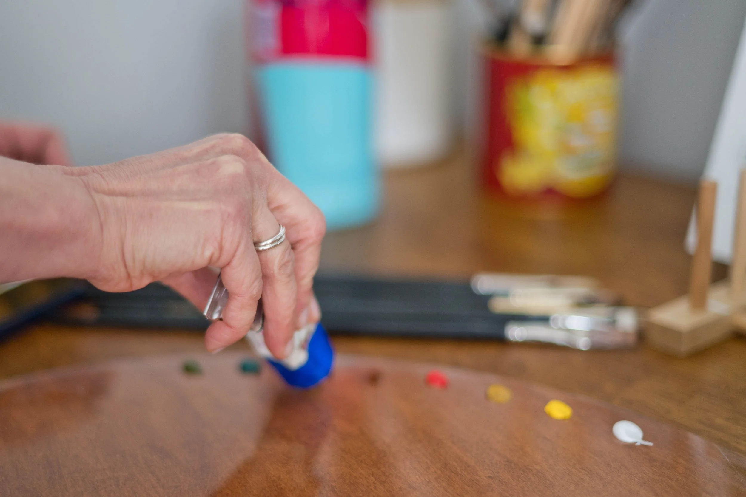 A person's hand holding a blue paint cap, with a wooden palette and small blobs of colorful paint on a table. In the background, there are containers with art supplies and paintbrushes.