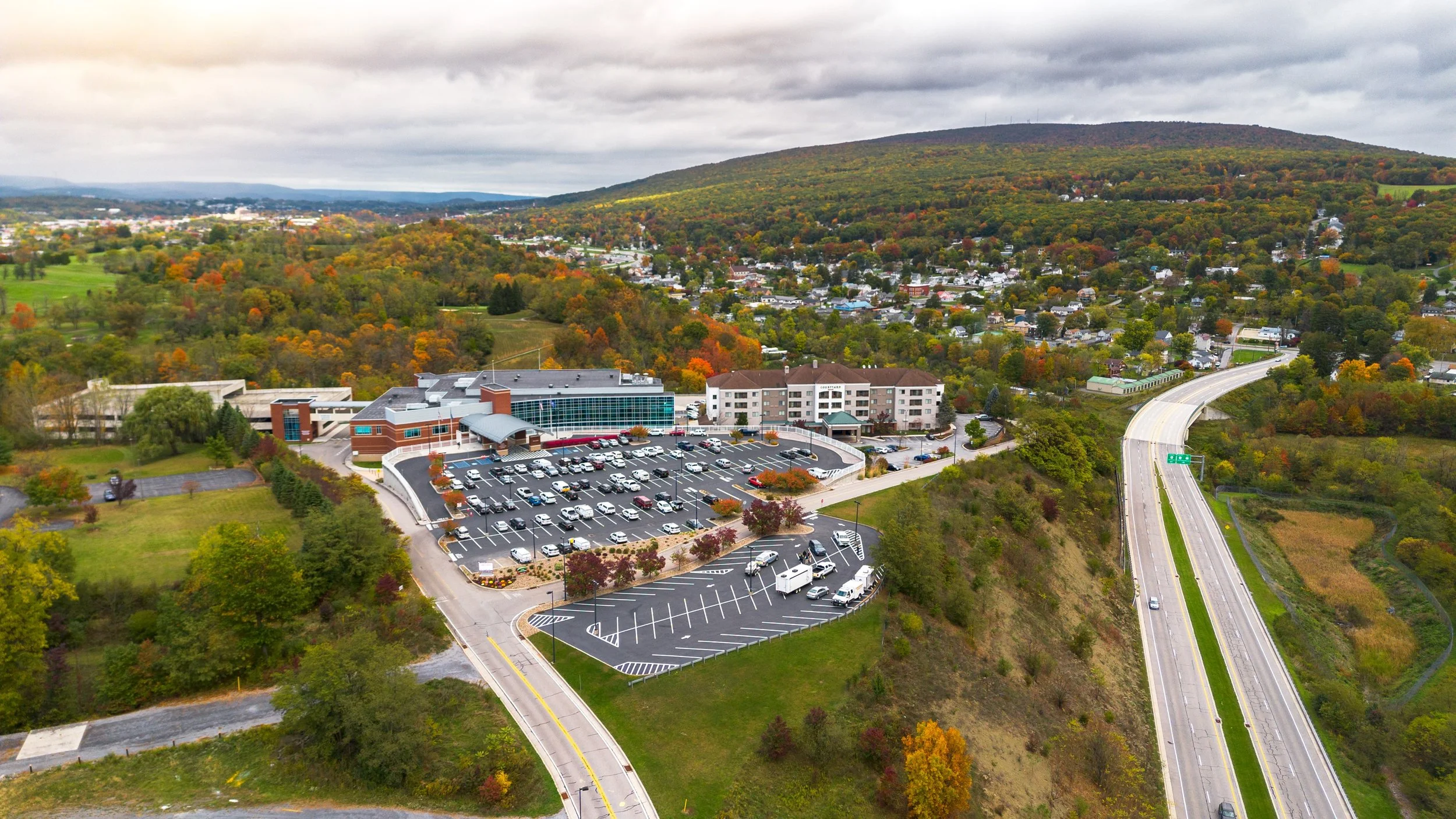 Aerial drone view of the Blair County Convention Center in Altoona, Pennsylvania, showcasing the facility's ample parking, adjacent hotel, and convenient highway access, surrounded by fall foliage across the Allegheny Mountains.