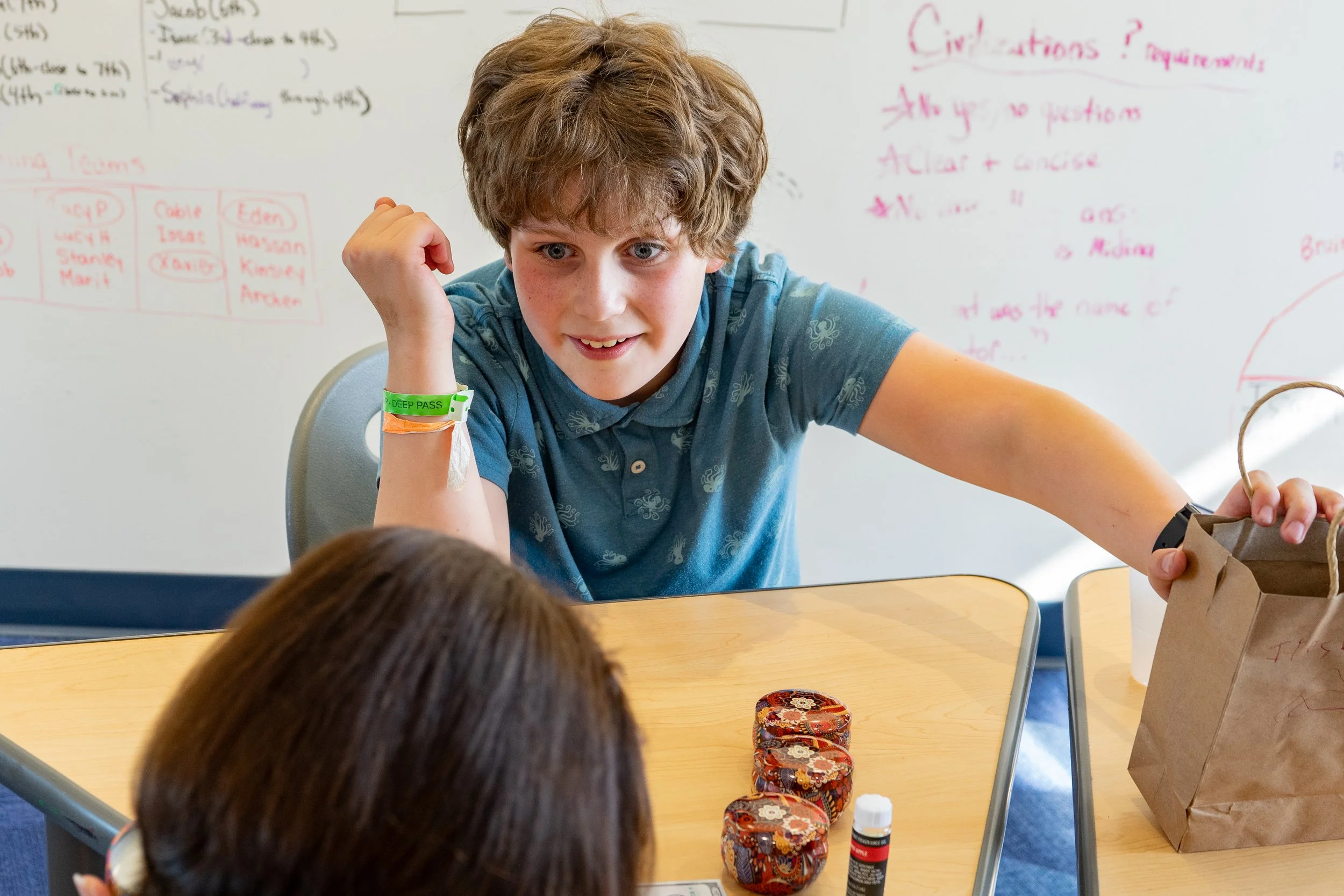 A teenage boy with curly hair and a green wristband smiles while reaching for a paper bag on a classroom table, with a girl with brown hair in front of him. The table has small jars and a glue stick. Whiteboard in the background has handwritten notes.