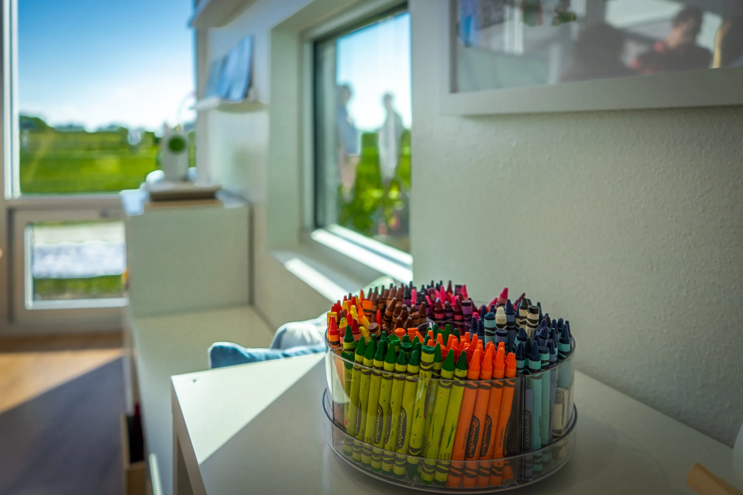 A clear plastic container filled with colorful Crayola crayons sits on a white surface in a room with large windows showing a sunny outdoor landscape.