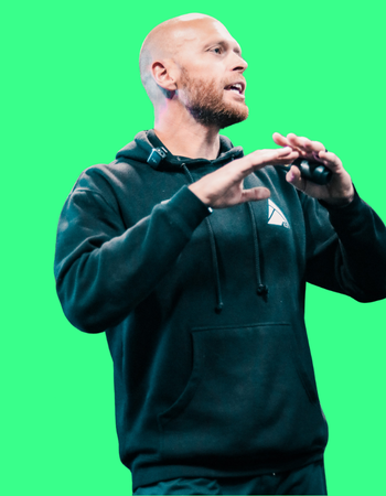 A bald man with a beard wearing a black hoodie, speaking or explaining something, with a green screen background.