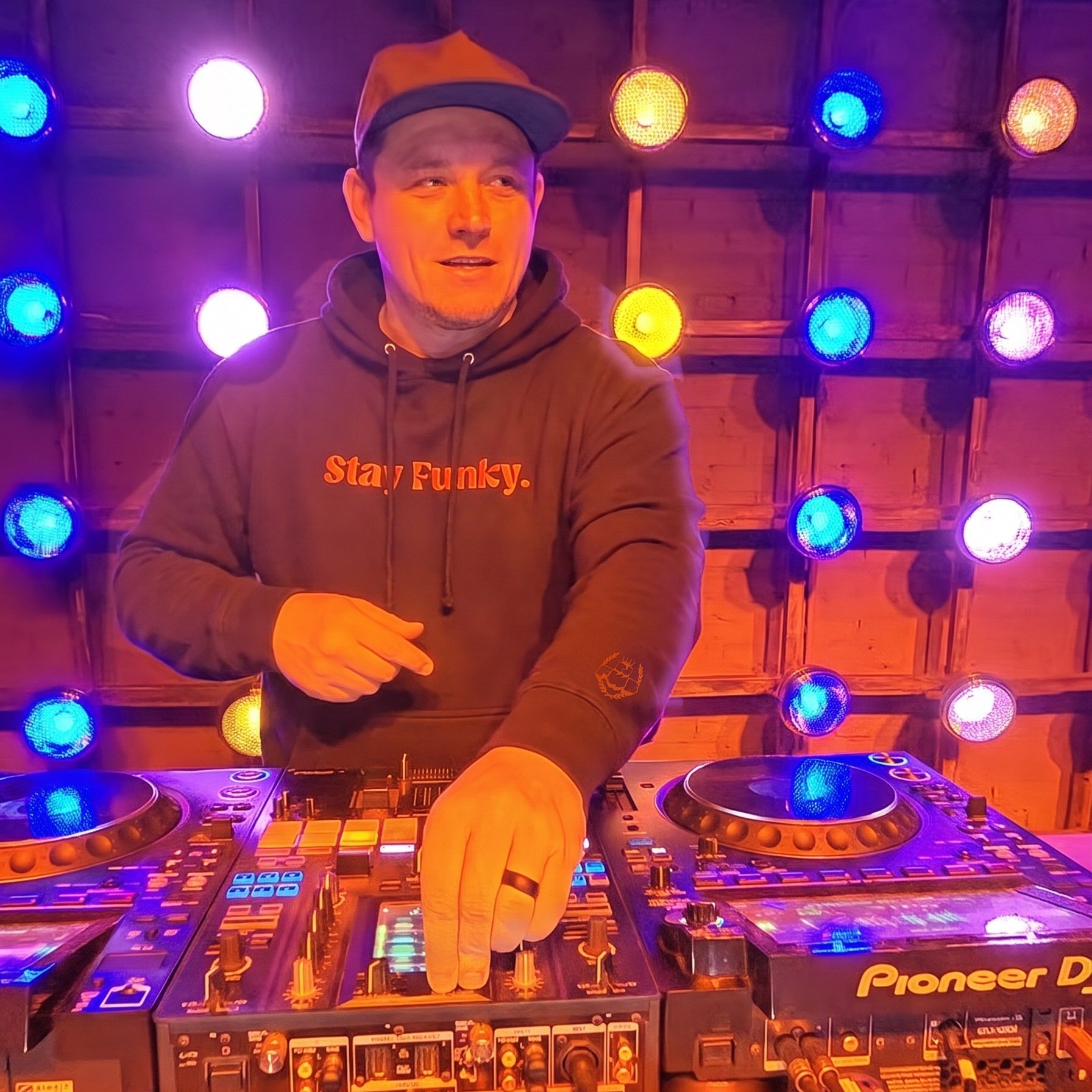 DJ FWB behind the decks wearing the black Secret Stash Heavyweight Hoodie with orange Stay Funky chest embroidery and the crest on the left wrist