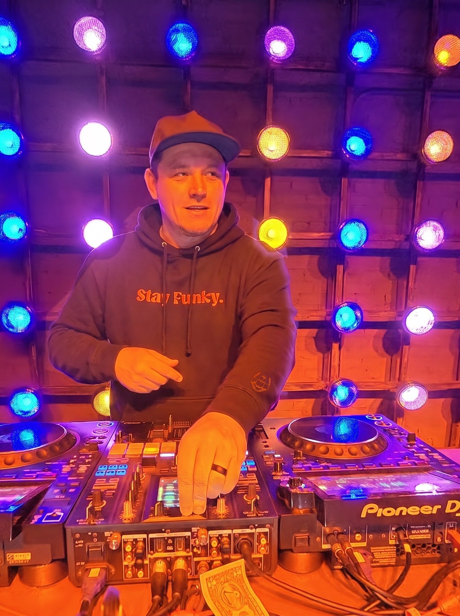 DJ FWB behind the decks in the black Secret Stash Heavyweight Hoodie with orange Stay Funky embroidery