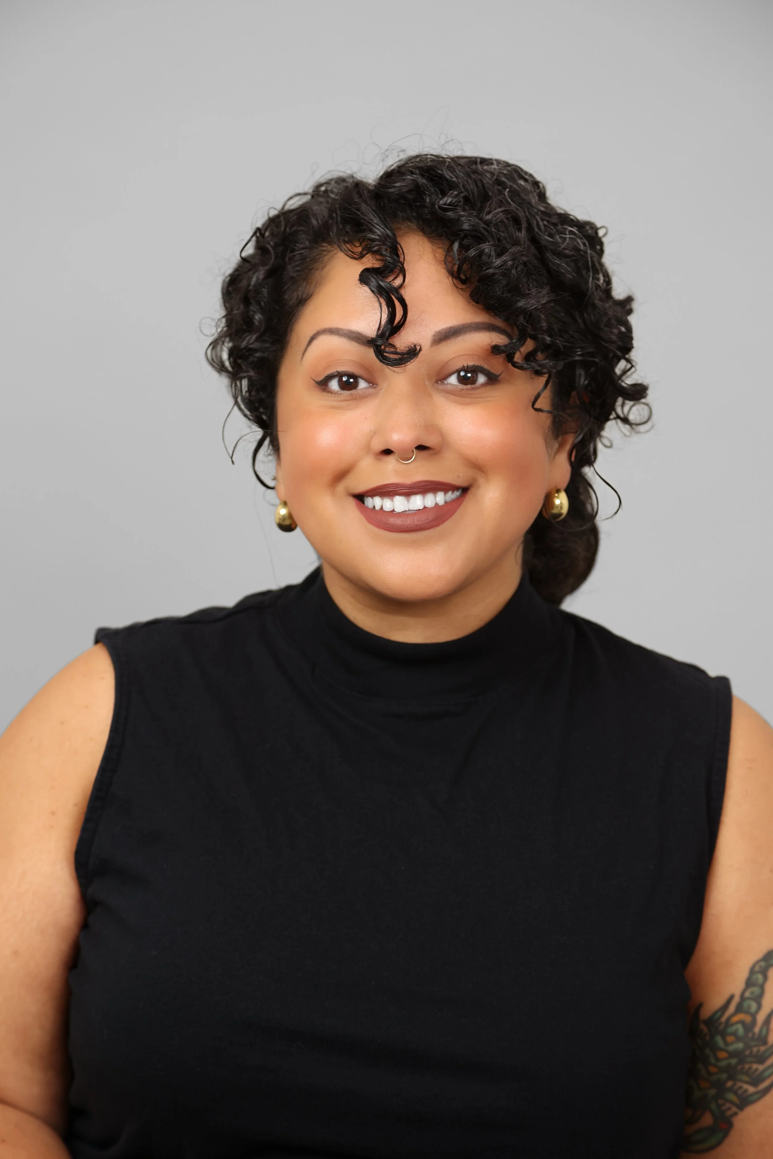 Close-up of a smiling woman with curly dark hair, wearing gold earrings, a septum piercing, and a sleeveless black top, against a plain gray background.