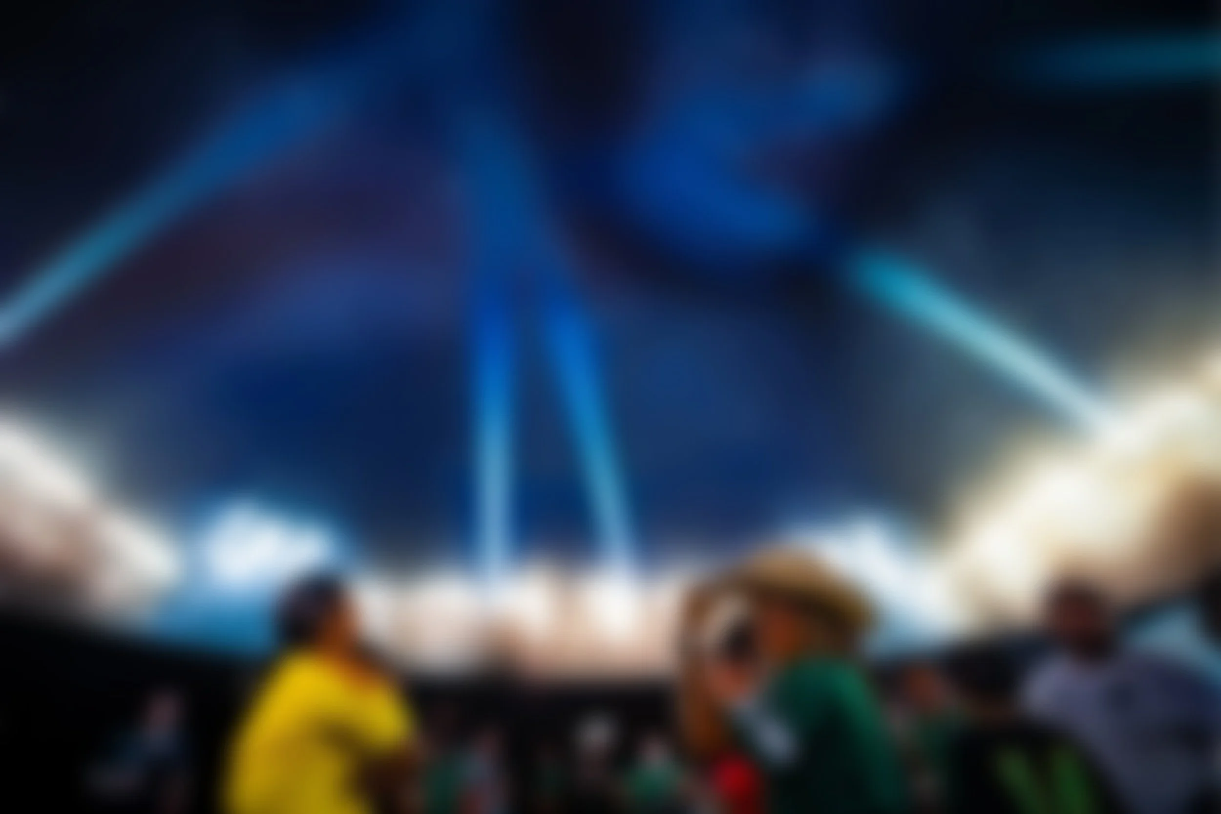 Blurred image of what appears to be a vibrant, colorful scene with lights and possibly people or objects, but specific details are indistinct due to the heavy blur.