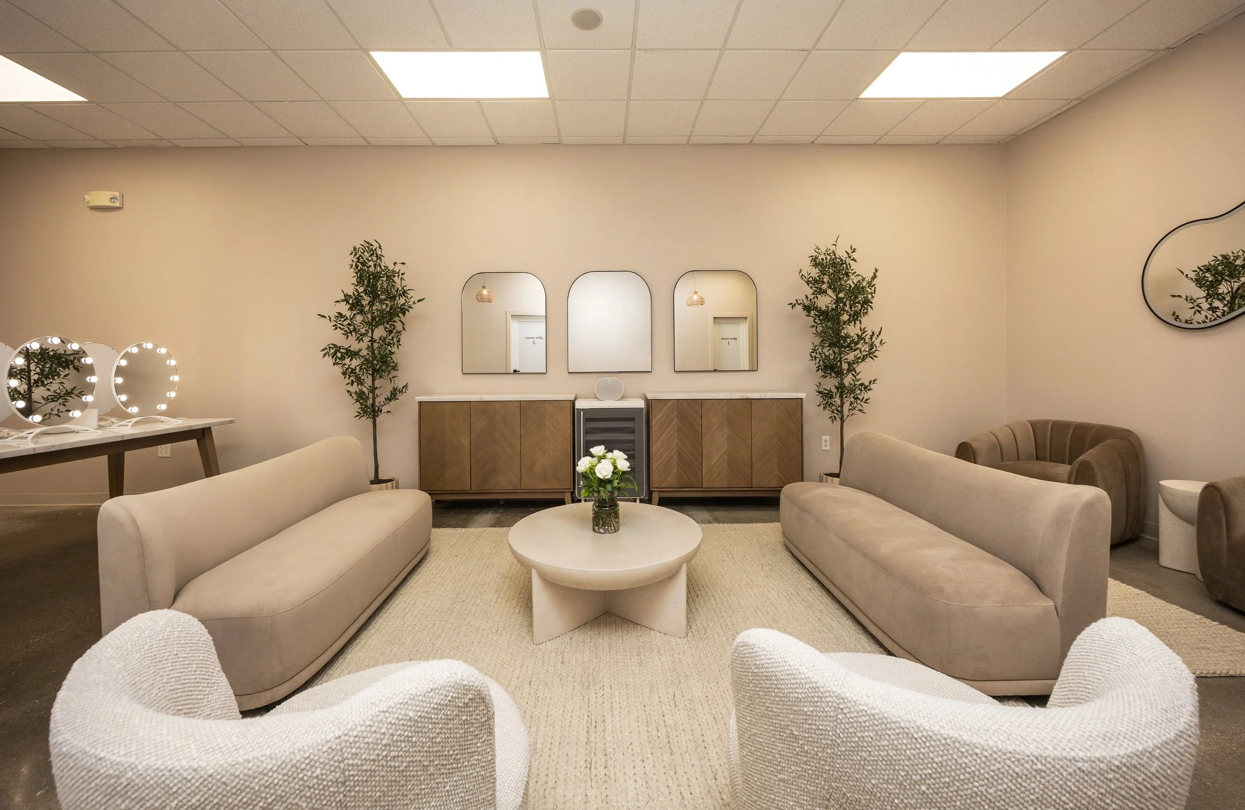 A modern, minimalistic lounge area with beige sofas, white textured armchairs, a round coffee table with a floral arrangement, two tall potted plants, a wooden sideboard with three mirrors, and decorative lighting.