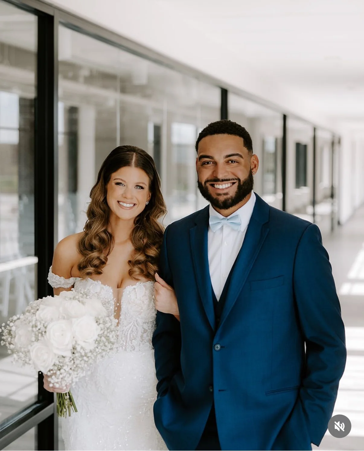 A smiling bride in a wedding dress holding a bouquet and a smiling groom in a dark blue suit with a white bow tie standing in a modern building corridor.