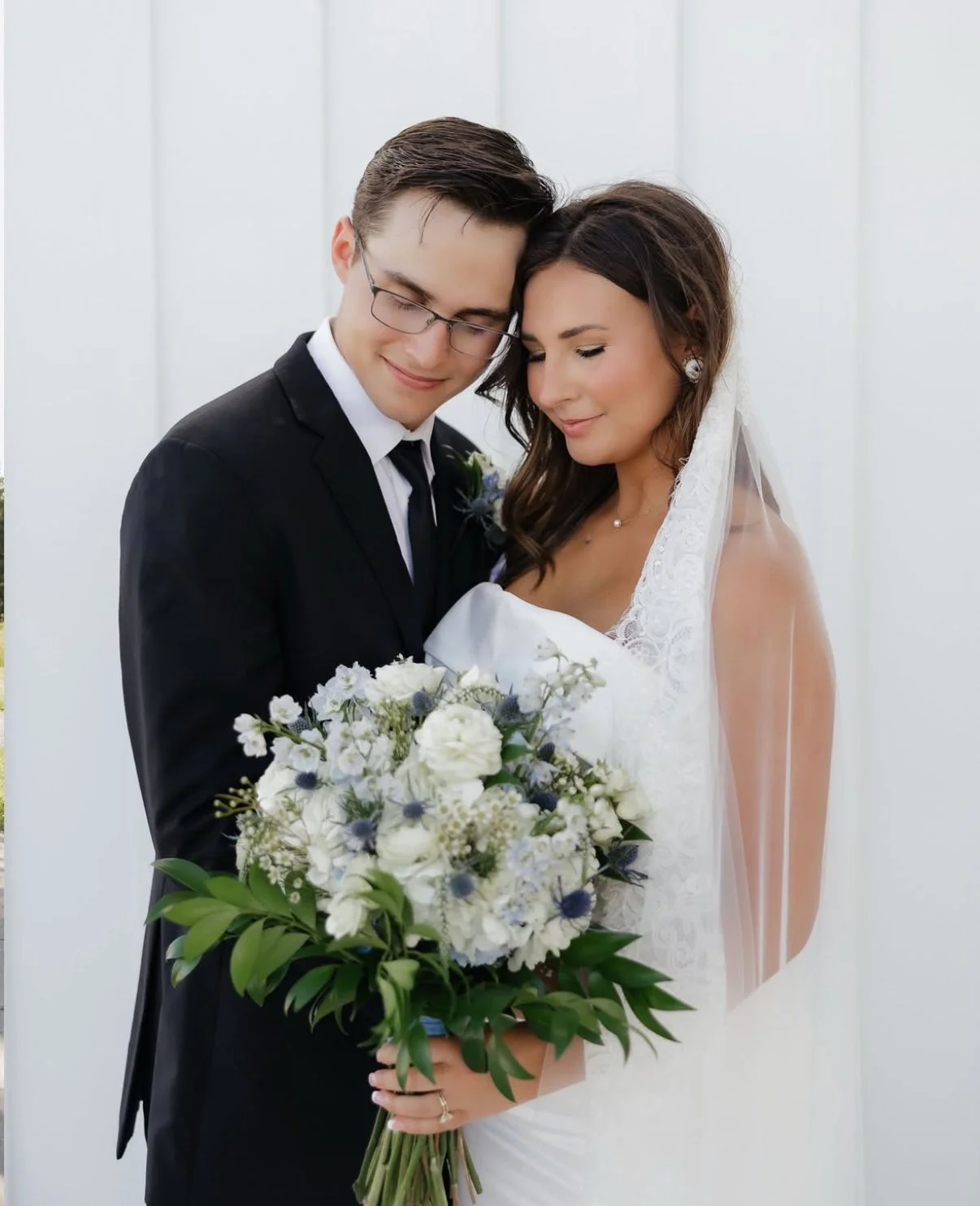 A bride and groom standing close together, holding a bouquet of white flowers, with their eyes closed and smiling.