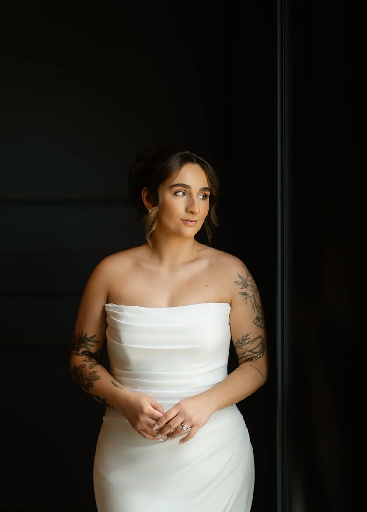 A woman in a strapless white dress with tattoos on her arms, standing by a window with soft natural light, looking thoughtfully to the side.