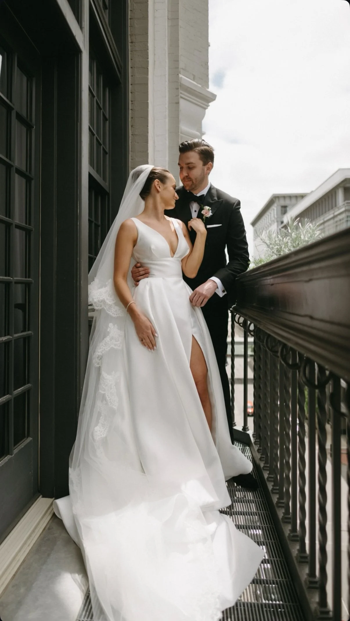 Bride and groom standing on a balcony, embracing each other. The bride is wearing a white wedding gown with a slit, veil, and jewelry; the groom is in a black tuxedo with a boutonniere. They are looking at each other affectionately.