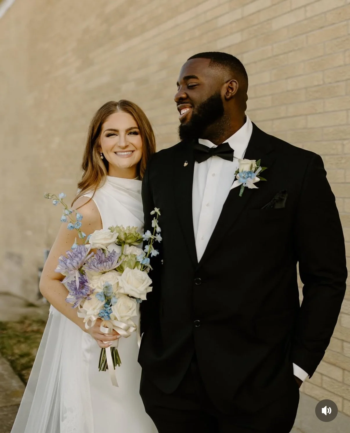 A bride and groom standing outdoors, smiling and dressed in wedding attire, with the bride holding a bouquet of flowers.