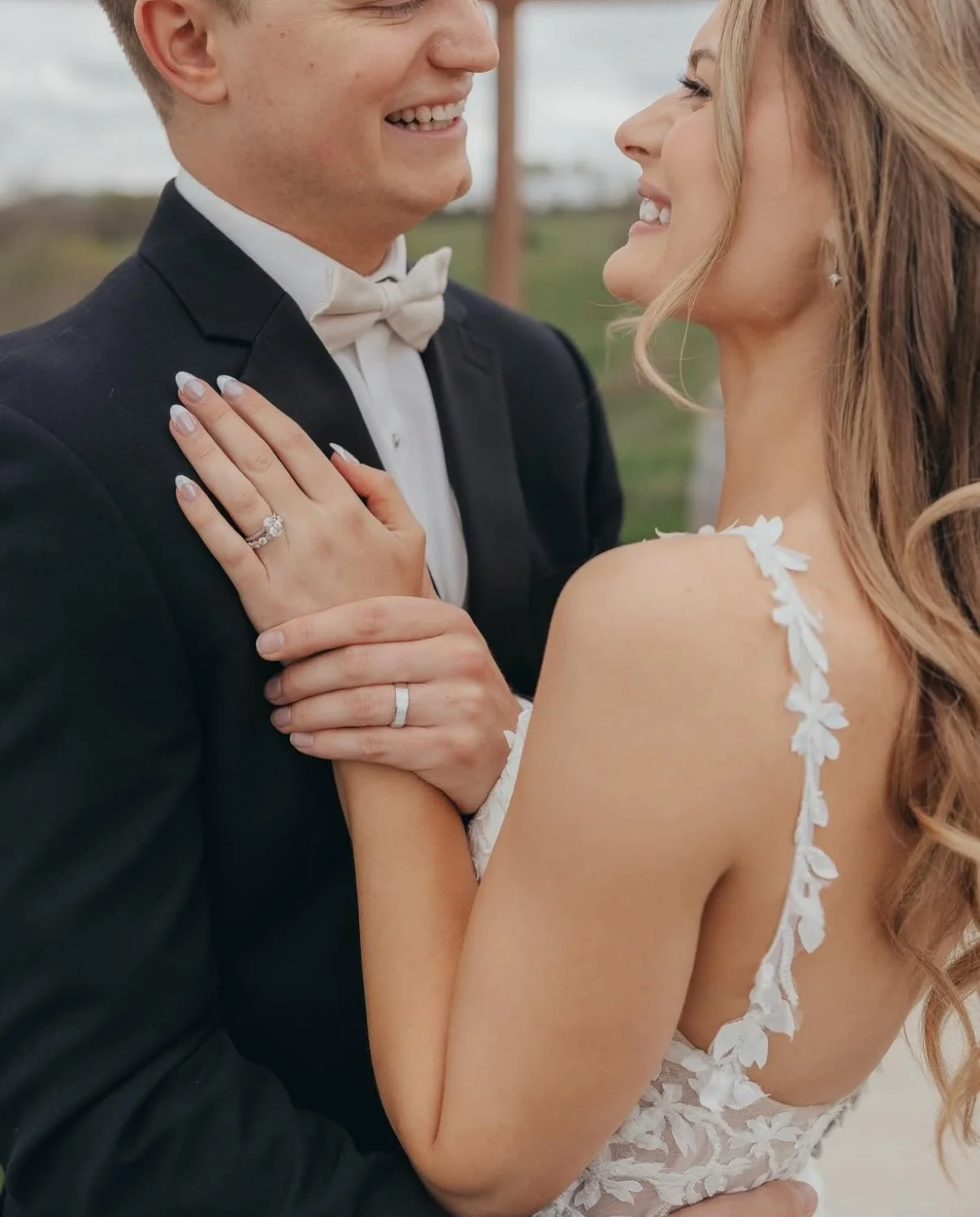 A bride and groom smiling and looking at each other, showing their wedding rings, with the bride wearing a white dress with floral details on the straps.