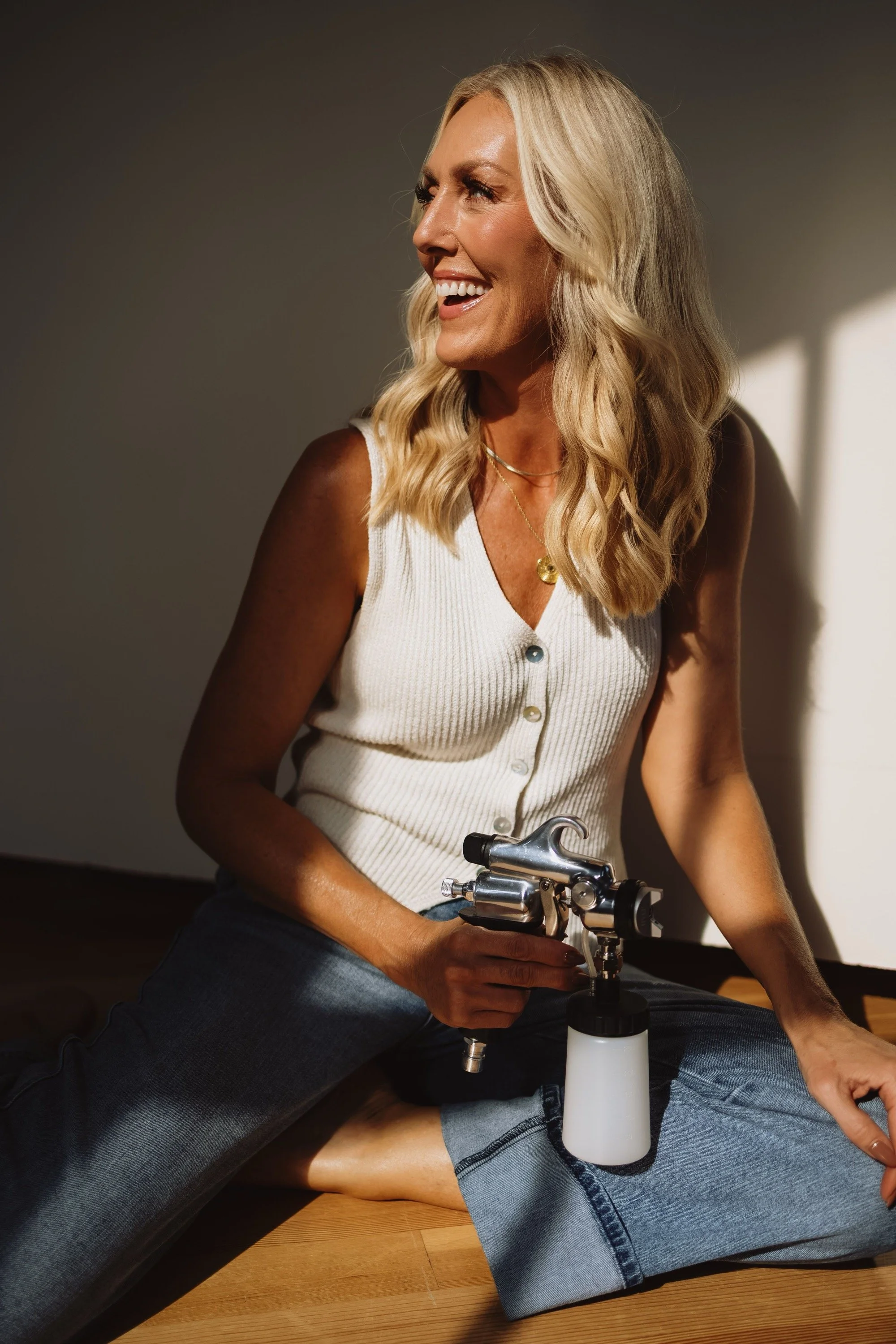 A blonde woman sitting on the floor, smiling, holding a spray tan gun.