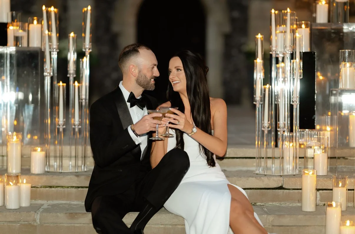 A man and woman in formal attire sitting on steps, holding glasses, surrounded by lit candles, at what appears to be a romantic evening or celebration.