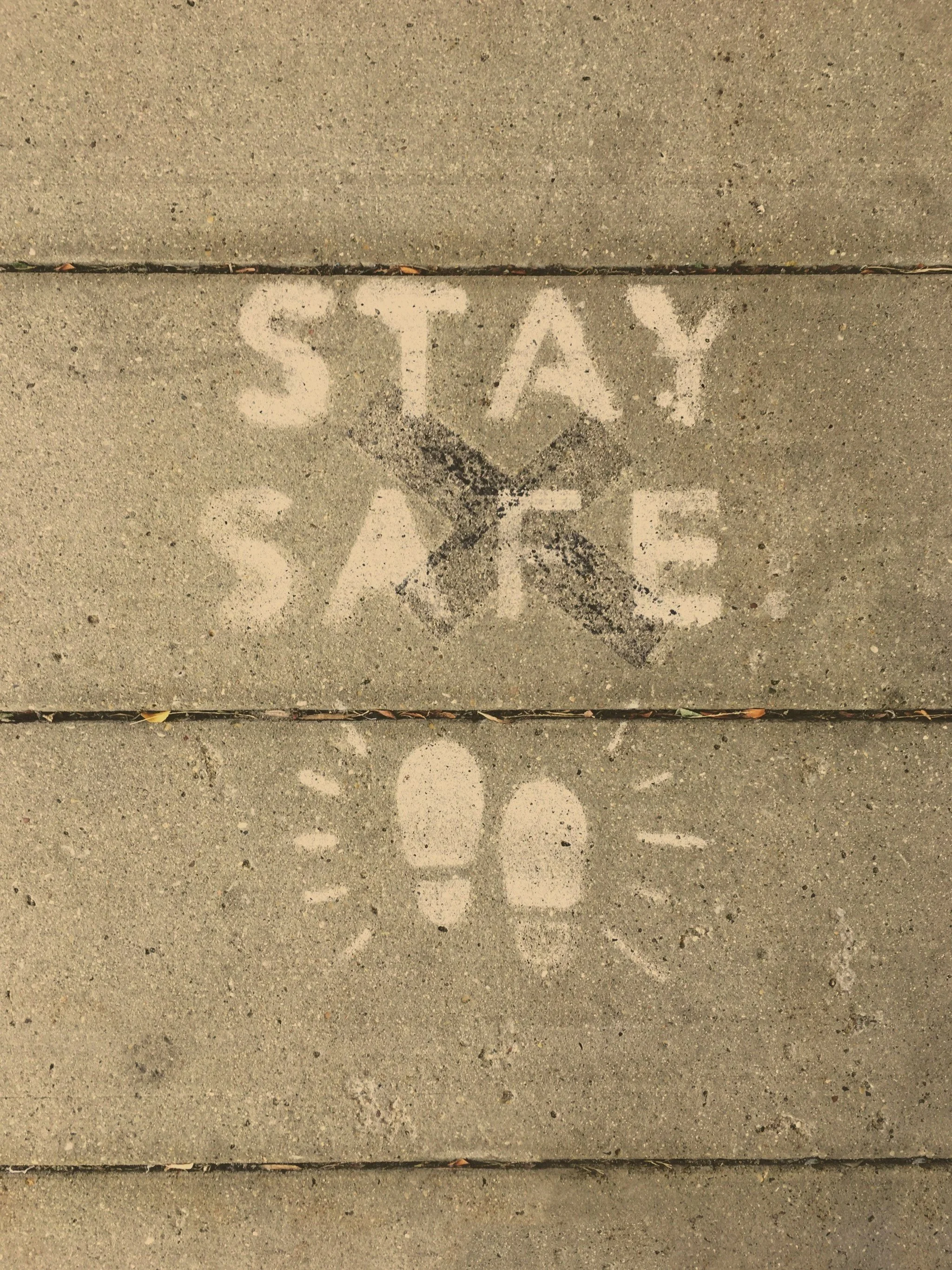 White sidewalk chalk markings on concrete sidewalk reading "STAY SAFE" above a footprint with radiating lines.
