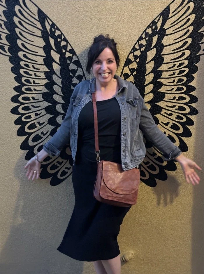Mara Adelman standing in front of a butterfly wing mural, smiling, wearing a black dress, denim jacket, and a brown crossbody bag.