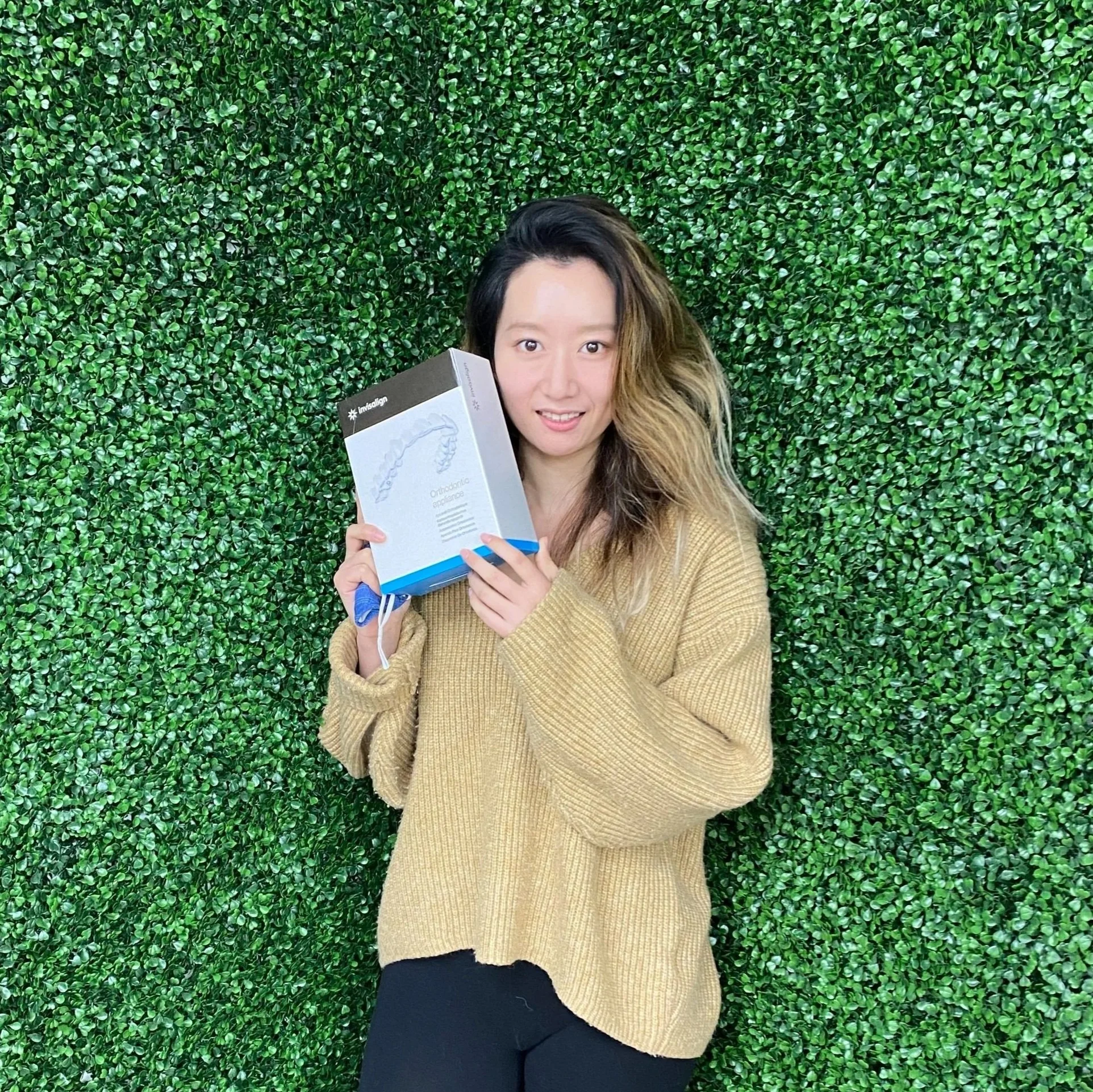 Young woman with long hair holding a box of Invisalign orthodontic aligners standing in front of a green leafy wall.