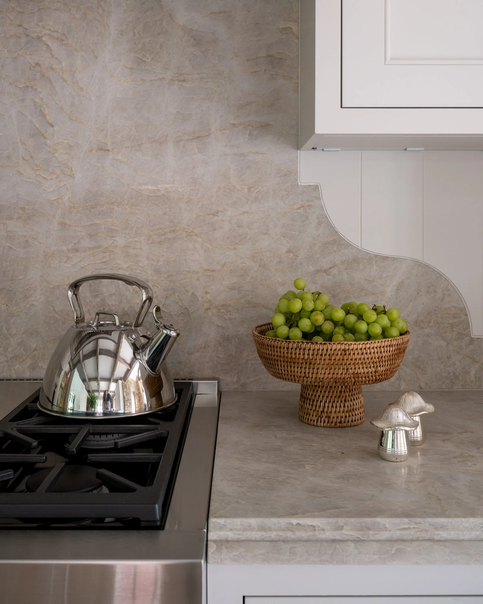 A kitchen counter with a stainless steel kettle on a stove, a woven basket of green grapes, and decorative mushroom-shaped objects, with white cabinetry and beige marble backsplash.