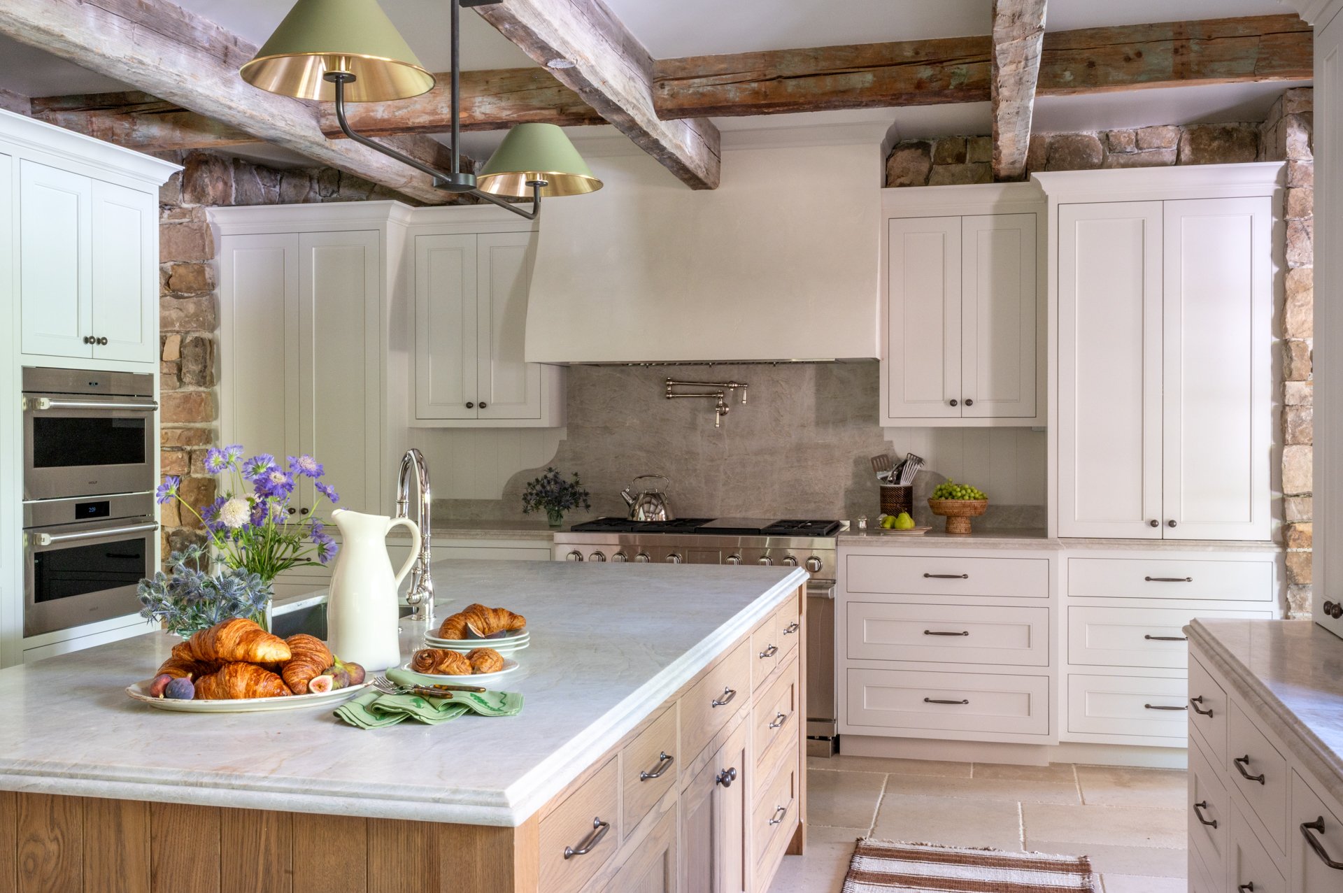 A kitchen with white cabinets, wooden accents, a gas stove, and a marble island. There is a bouquet of flowers, croissants, and a pitcher on the island, with exposed wooden beams on the ceiling.