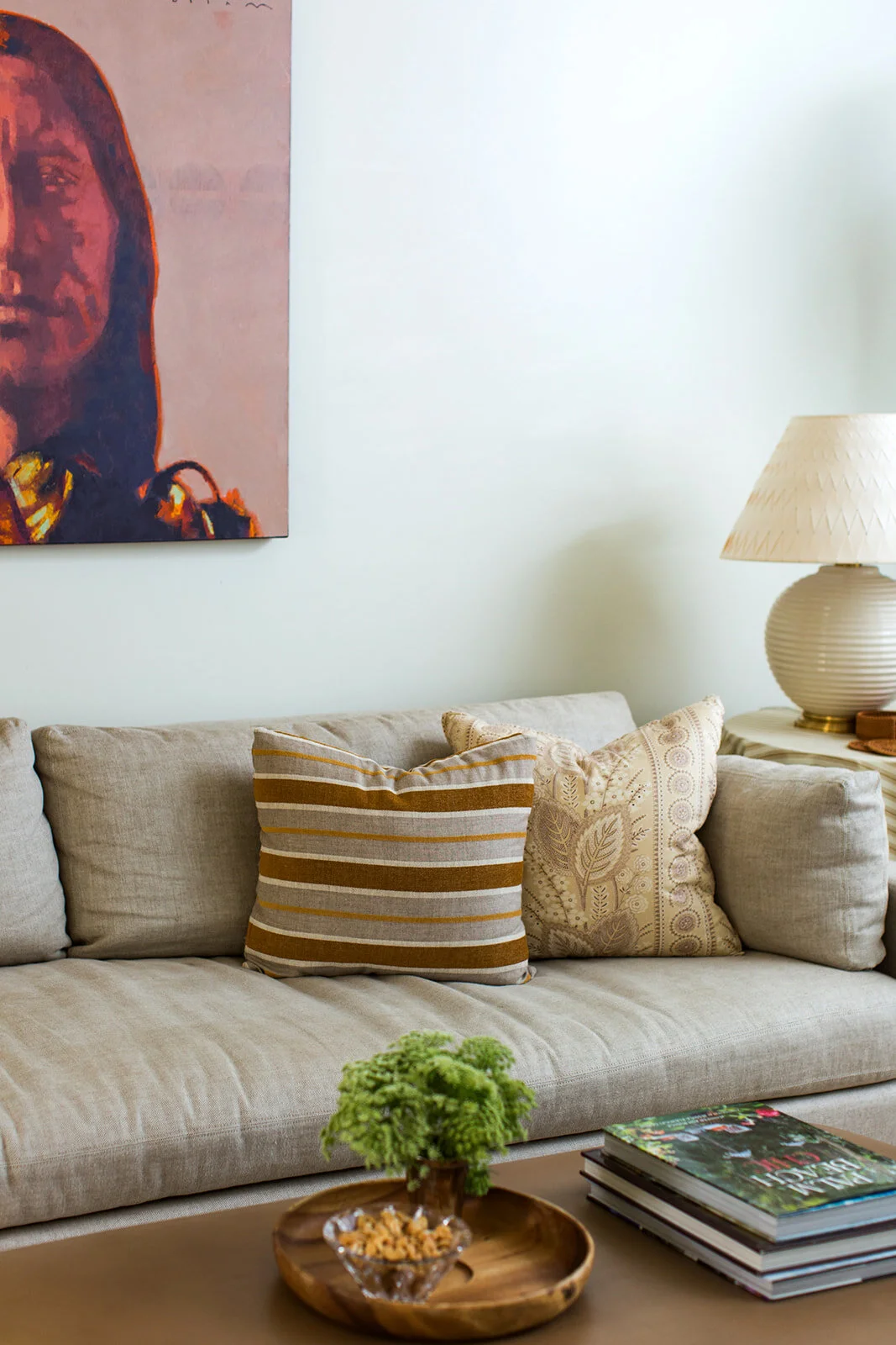A beige sofa with two decorative pillows, a wooden coffee table with a small potted green plant and a bowl of snacks, a side table with a textured white lamp, and a large portrait of a woman hanging on the light-colored wall.