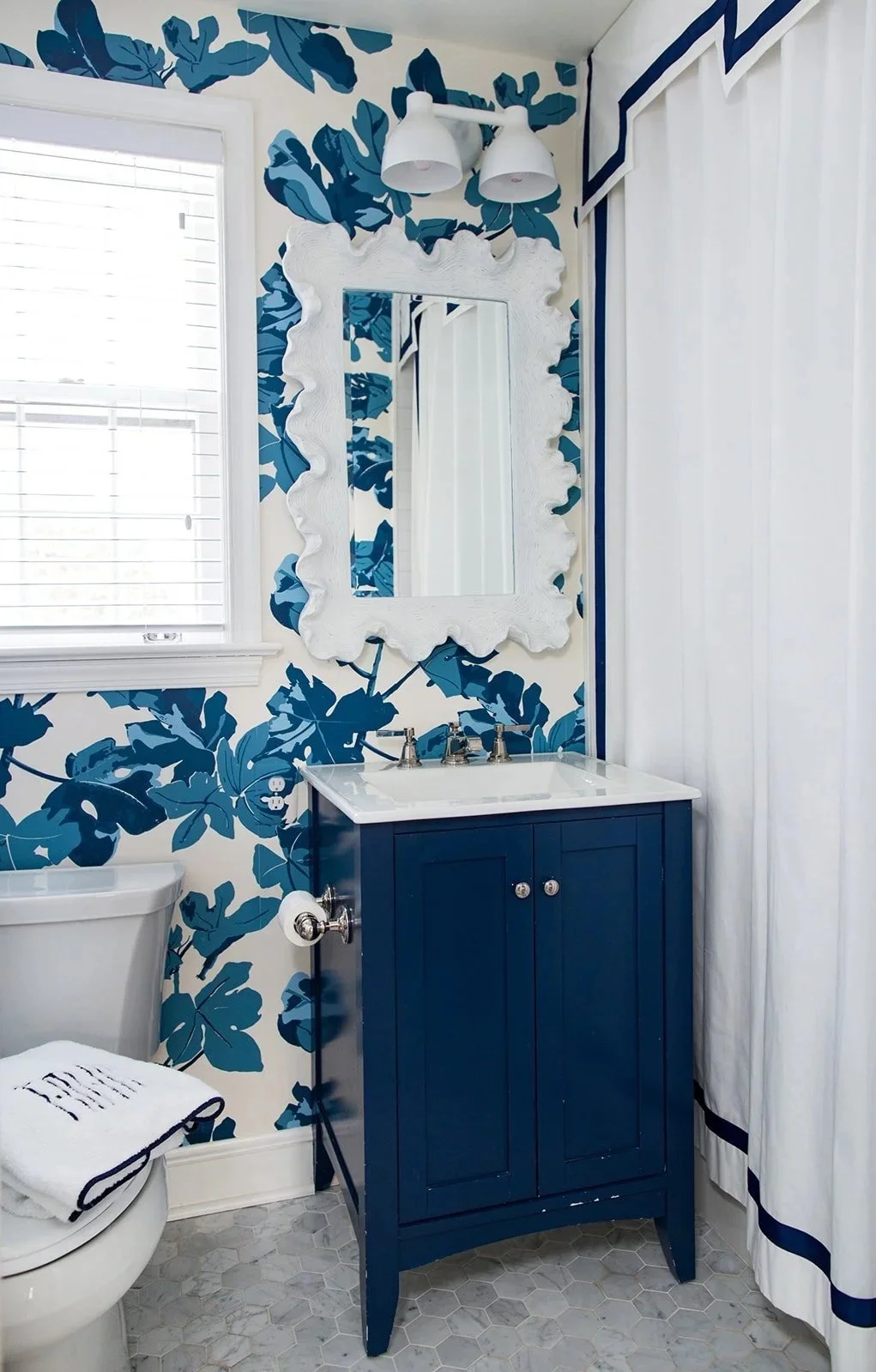 A bathroom with blue patterned wallpaper, a white window with blinds, a white-framed mirror, a navy blue vanity with a white sink, a white toilet with a towel on it, and a shower curtain with a blue border.