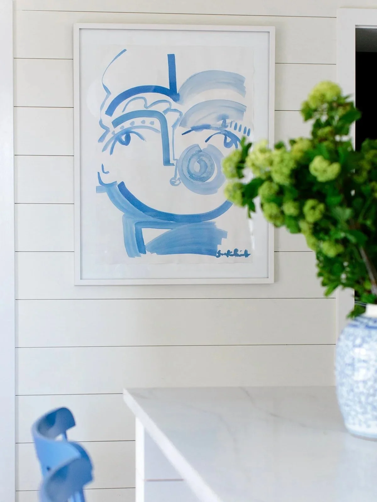 Abstract blue line drawing of a face and a nose, framed and hung on a white shiplap wall, with a green plant in a blue vase in the foreground.