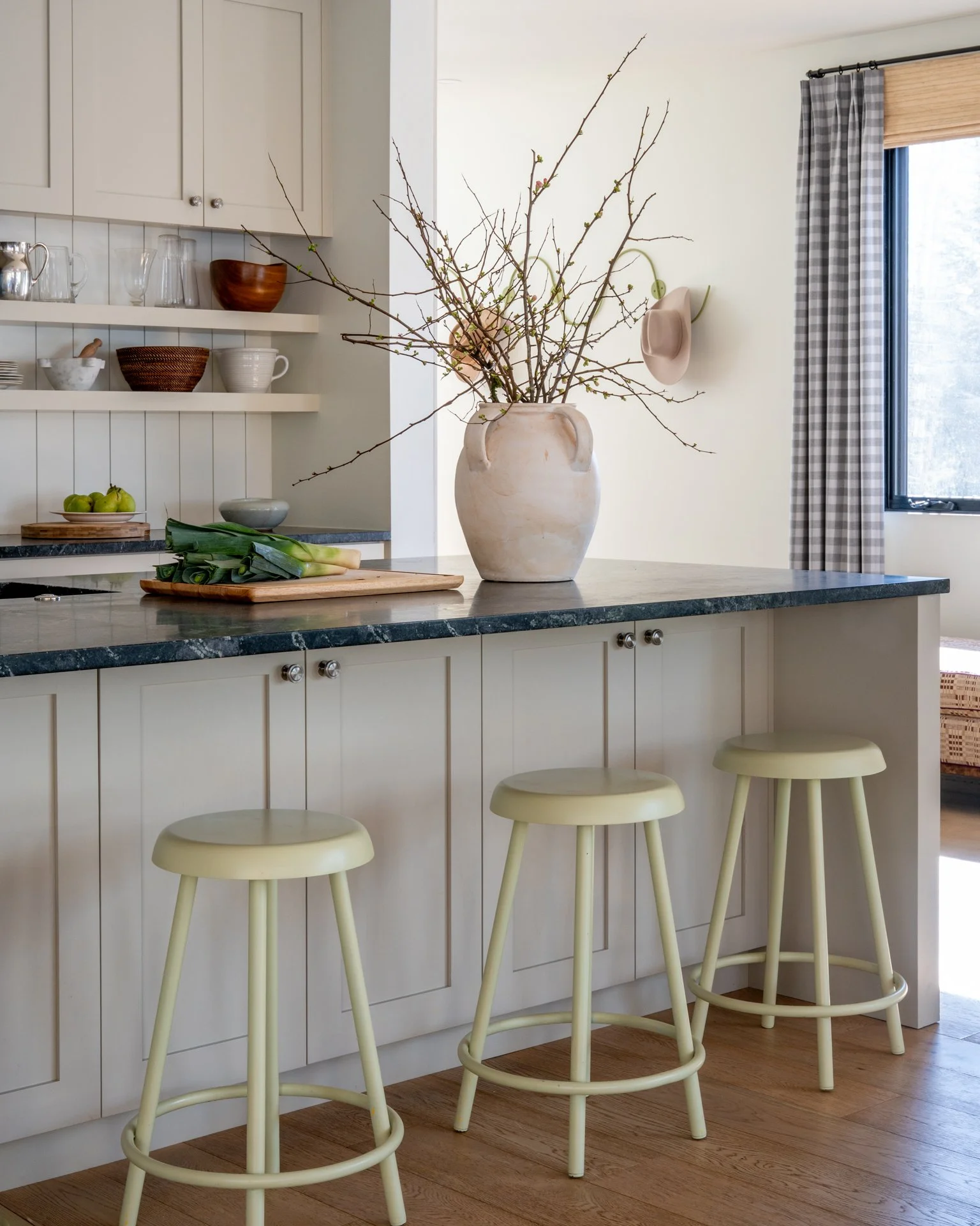 A kitchen with white cabinets, a dark green marble countertop, a large beige vase with branches on the counter, three pale yellow barstools, and a window with plaid curtains.