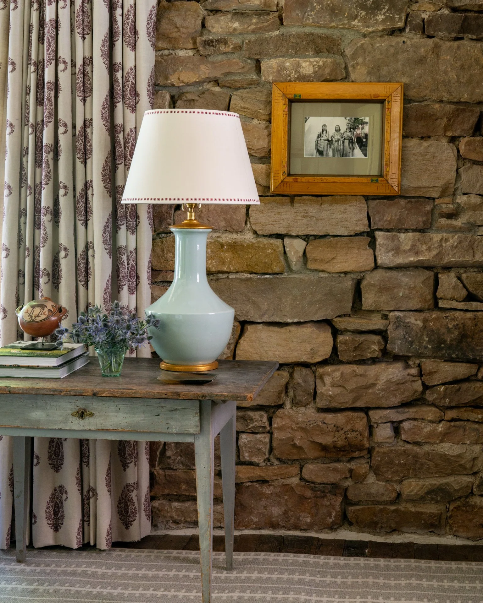 Interior scene with a rustic stone wall, a wooden table with a white and gold lamp, a small vase with purple flowers, a stack of books, and a framed black-and-white photo of three women in traditional dress. There are floral curtains to the left.