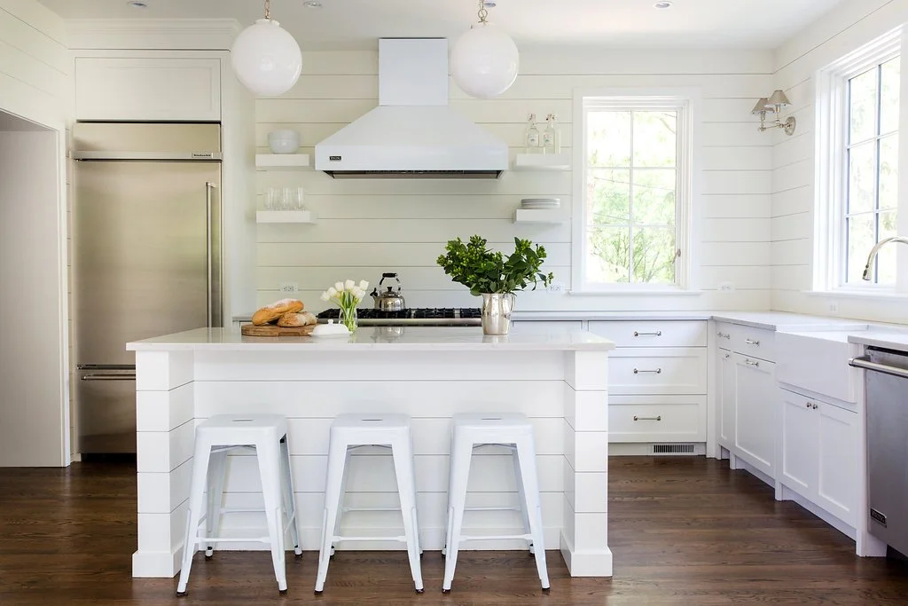 Bright kitchen with white cabinets, a white island with three white stools, a stainless steel refrigerator, and large windows letting in natural light. Decor includes a potted plant, a vase of flowers, and a bamboo tray with bread.