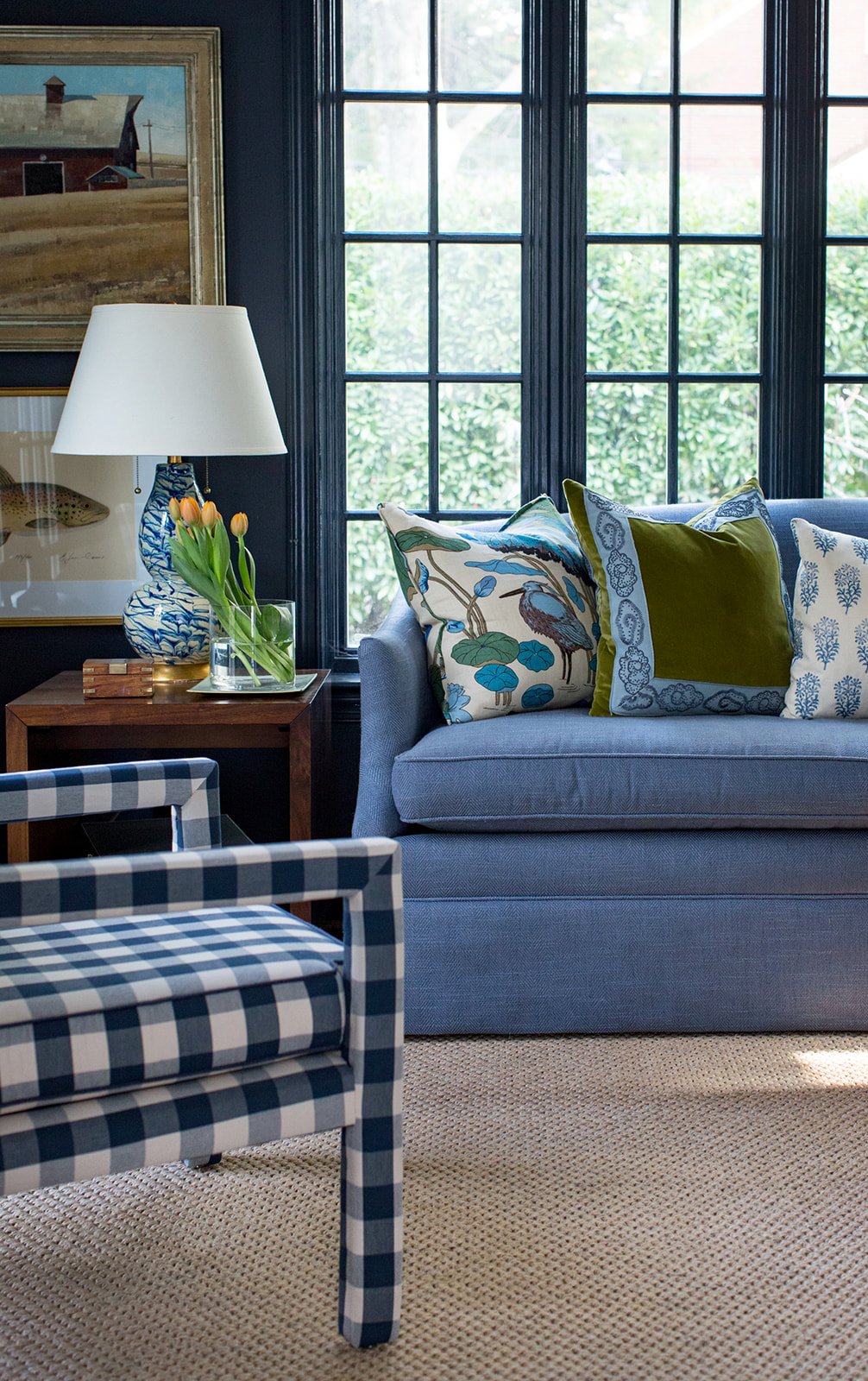 A cozy living room corner featuring a blue sofa with colorful patterned and velvet pillows, a wooden side table with a ceramic lamp, tulips in a glass vase, and an armchair with blue and white checkered upholstery, all set against large window panes 