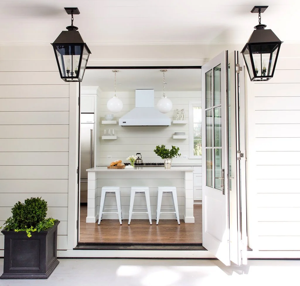 View from outside into a bright, white kitchen with a door open. The kitchen has white cabinets, a white island with three white bar stools, and hanging white spherical pendant lights. Two black outdoor lanterns are visible in the frame, and there is