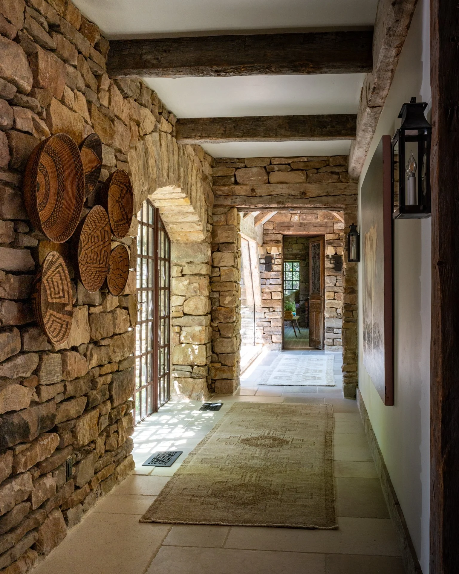 A hallway with stone walls, wooden beams on the ceiling, a window, and a door at the end, with a beige rug on the tiled floor.