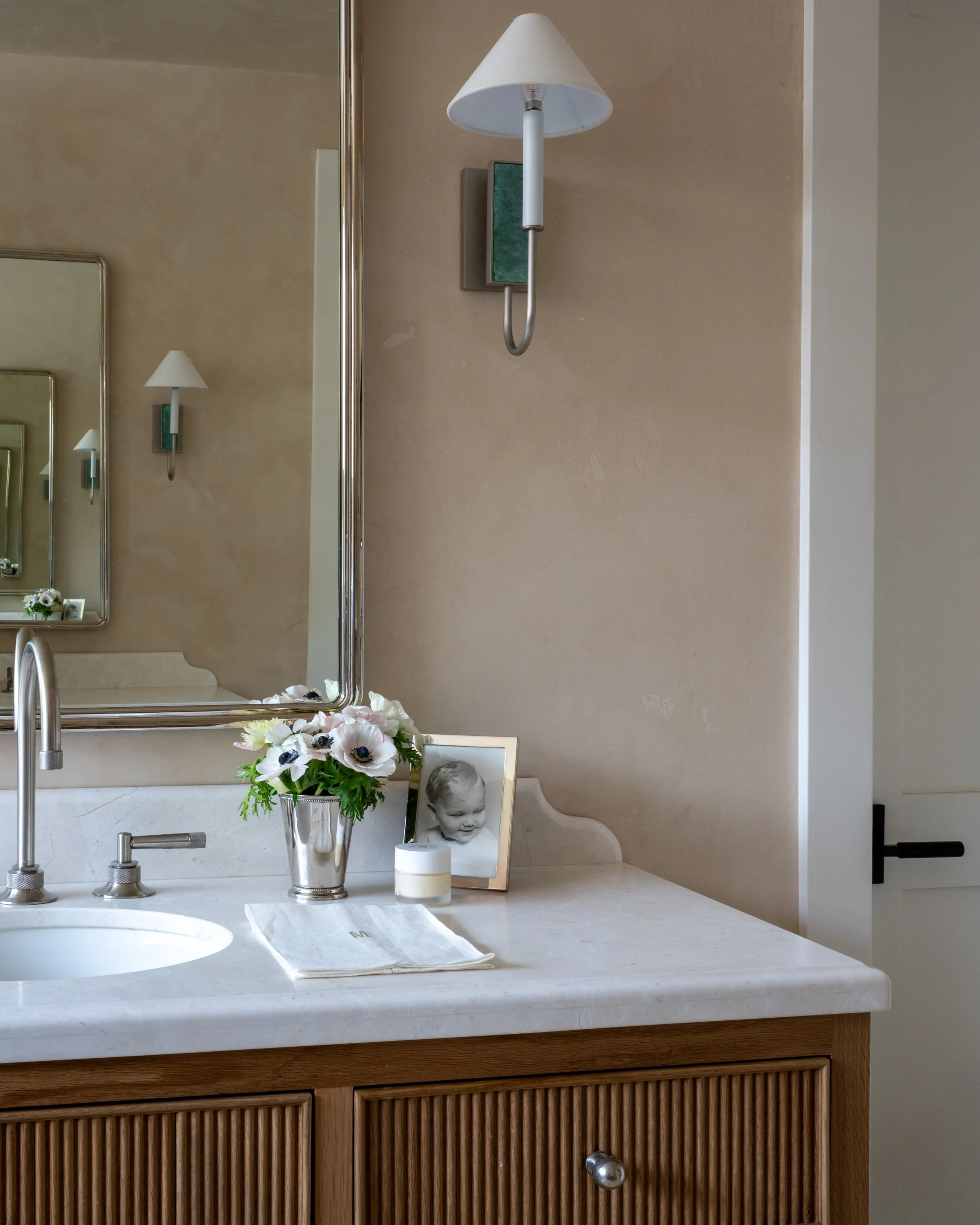 Bathroom vanity with a flower arrangement, a framed photo of a baby, a container, a white towel, a mirror, a sink, a faucet, and a wall-mounted lamp with a white shade.