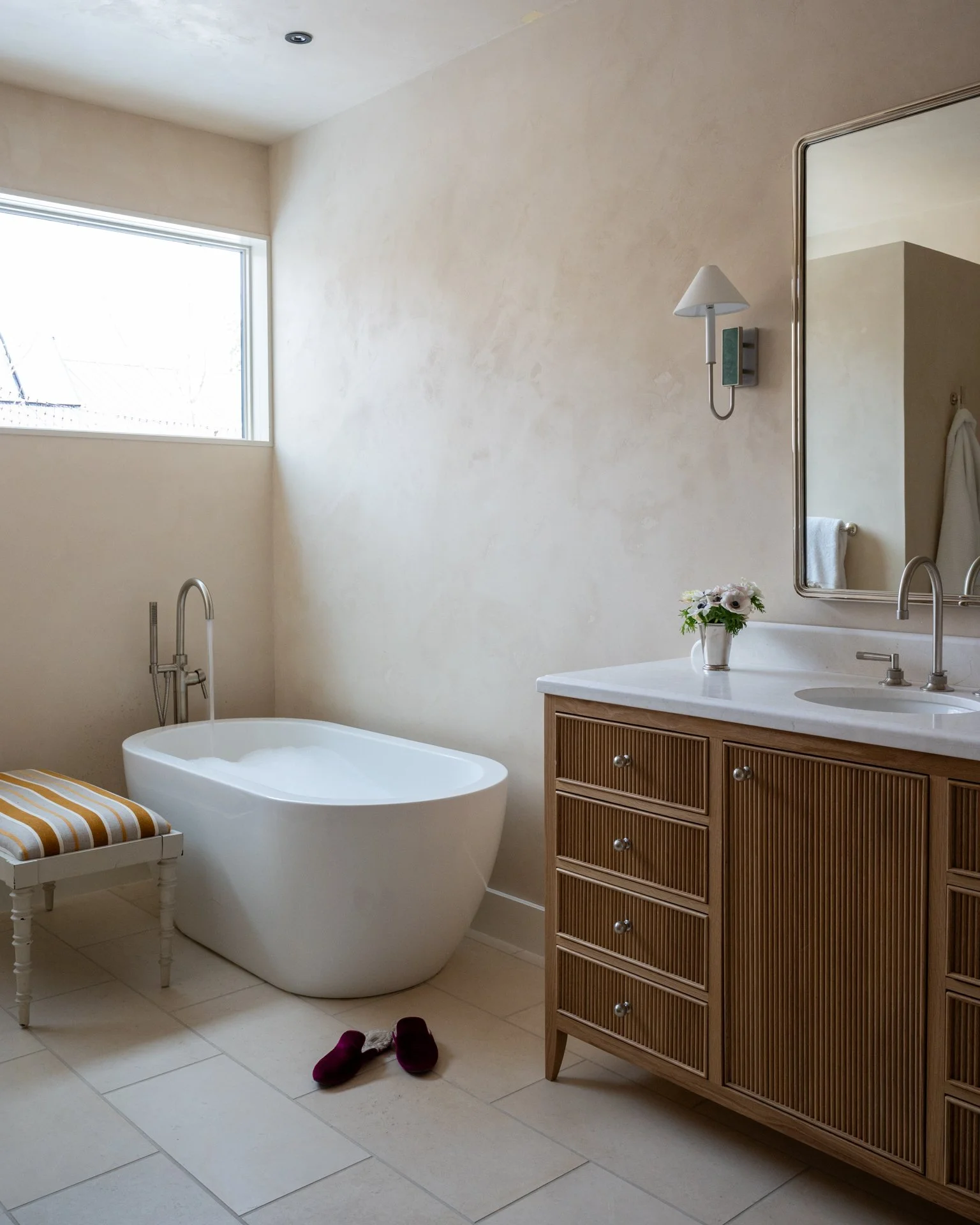 A bathroom with a standalone bathtub, a wooden vanity with a white countertop, a large mirror, a wall-mounted light fixture, a small white chair with a striped cushion, a vase with white flowers, and slippers on the floor.