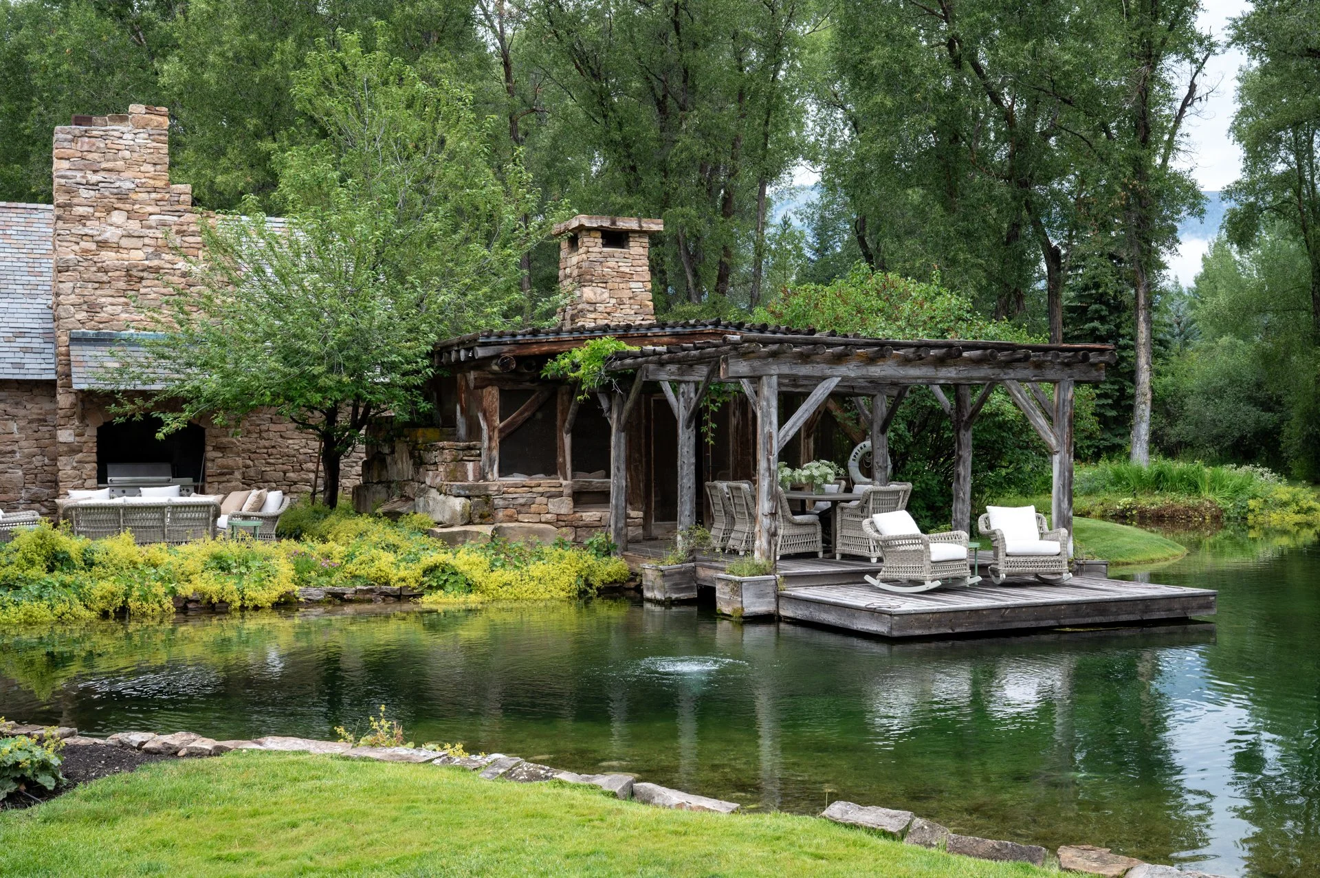 A riverside outdoor patio with seating and a wooden pergola surrounded by lush greenery and trees.