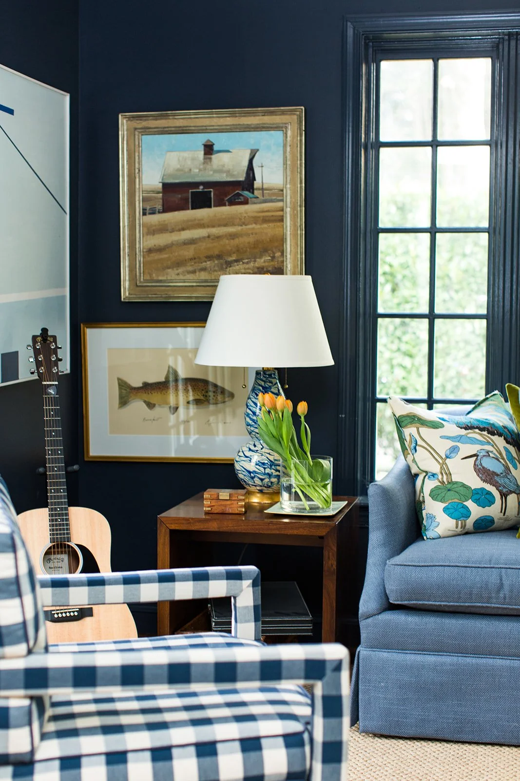 Cozy living room with dark blue walls, vintage barn painting, fish illustration, and large window; includes a table with tulips, a blue and white vase, a lamp, a gray sofa with bird-patterned pillows, and a guitar.