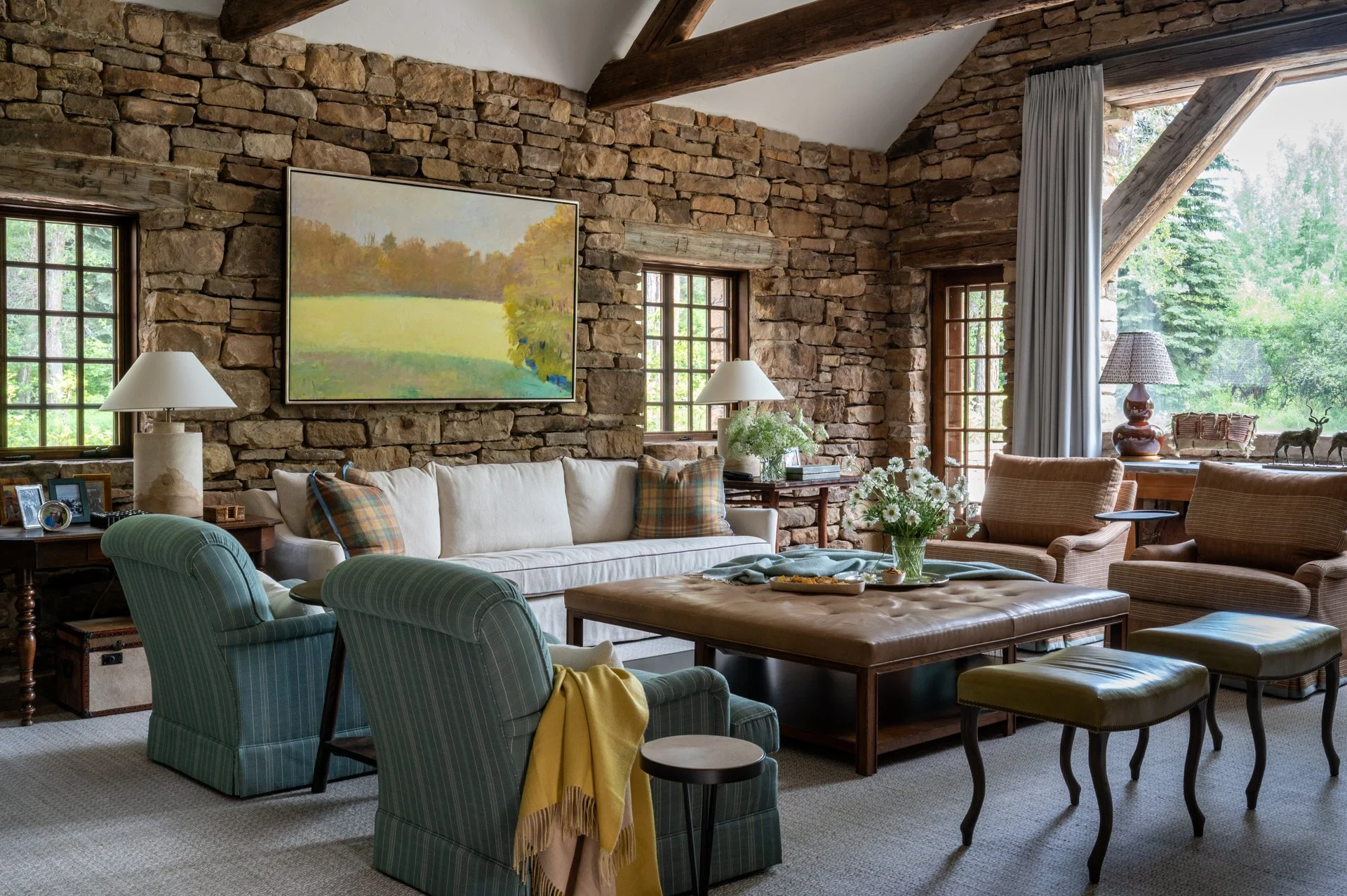 Living room with stone walls, wooden beams, large windows, and various seating including armchairs and a sofa, decorated with lamps, books, and art.
