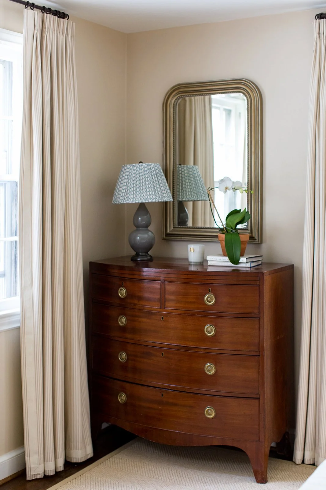 A wooden dresser with a polished finish and brass knobs, topped with a lamp, orchid plant, candle, and books, in a room with beige walls and cream curtains reflected in an oval mirror.