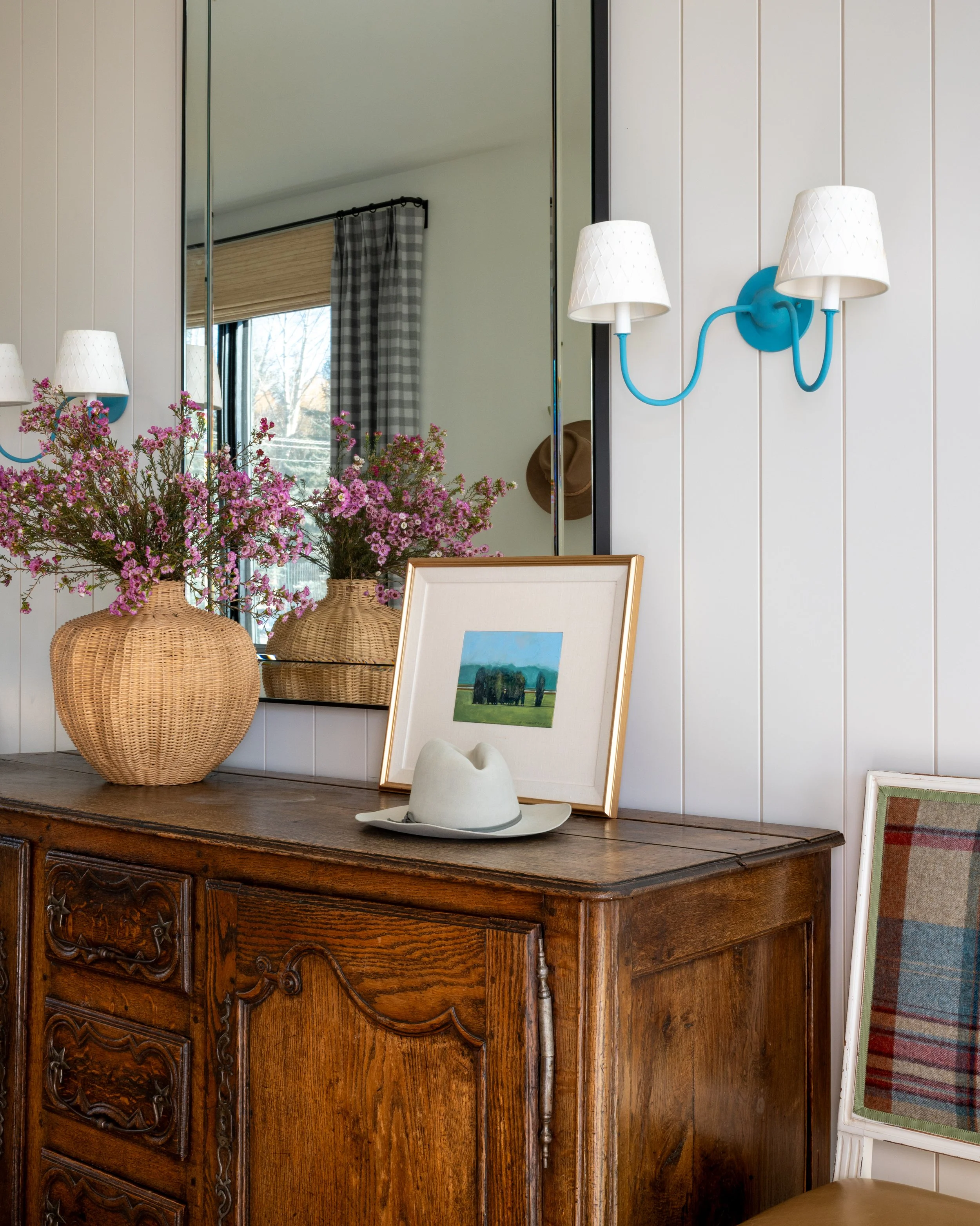 Room with a wooden dresser, framed painting, a white hat on a gray leaf bowl, and flowers in a basket. A mirror reveals a window with curtains and trees outside. Wall-mounted lamps with white shades and a blue backplate are also visible.