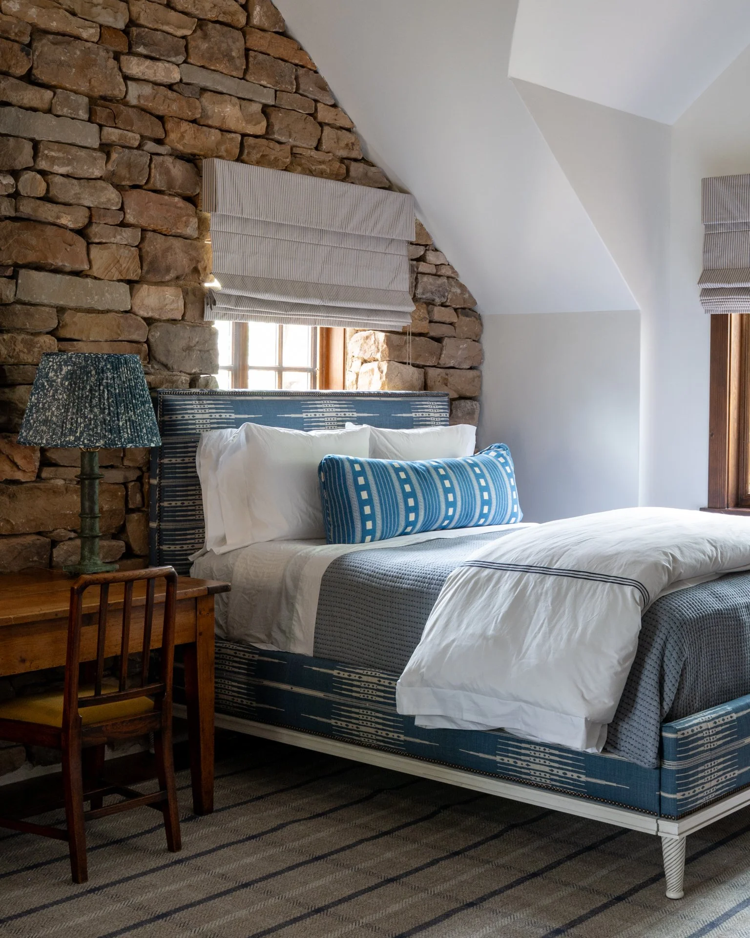 A bedroom featuring a stone wall with two windows, a bed with blue and white patterned bedding, a striped pillow, a small wooden desk, and a green bedside lamp with a floral lampshade. The room has white ceiling and walls with a sloped ceiling design