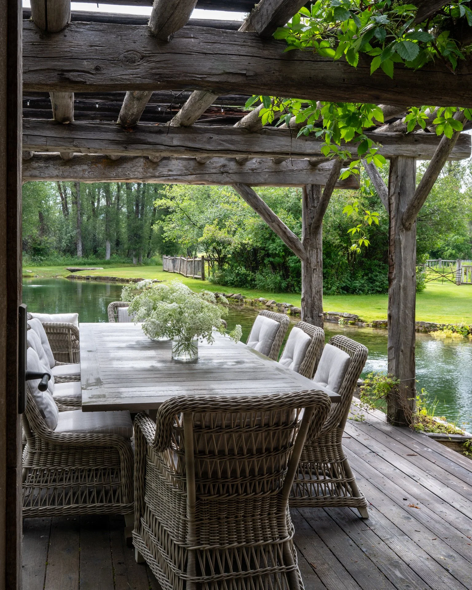 An outdoor dining area under a rustic wooden pergola overlooking a pond and lush green trees. The table is decorated with a vase of white flowers and surrounded by wicker chairs.