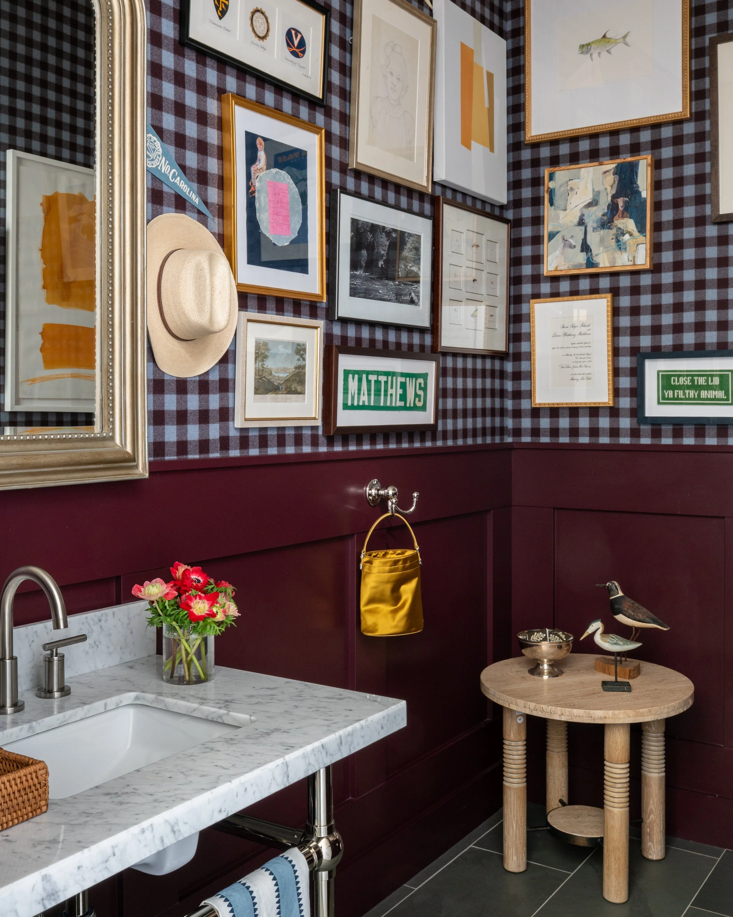 A kitchen with a white marble countertop, a sink, and a flower vase. The wall above features a gallery of framed pictures and artwork, including a fish painting, a portrait sketch, and various signs. A straw hat rests on the gallery frame, and a yell